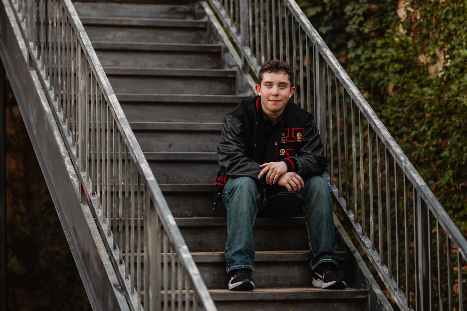 A person sits casually on concrete steps outdoors wearing jeans and a dark jacket in an urban setting.