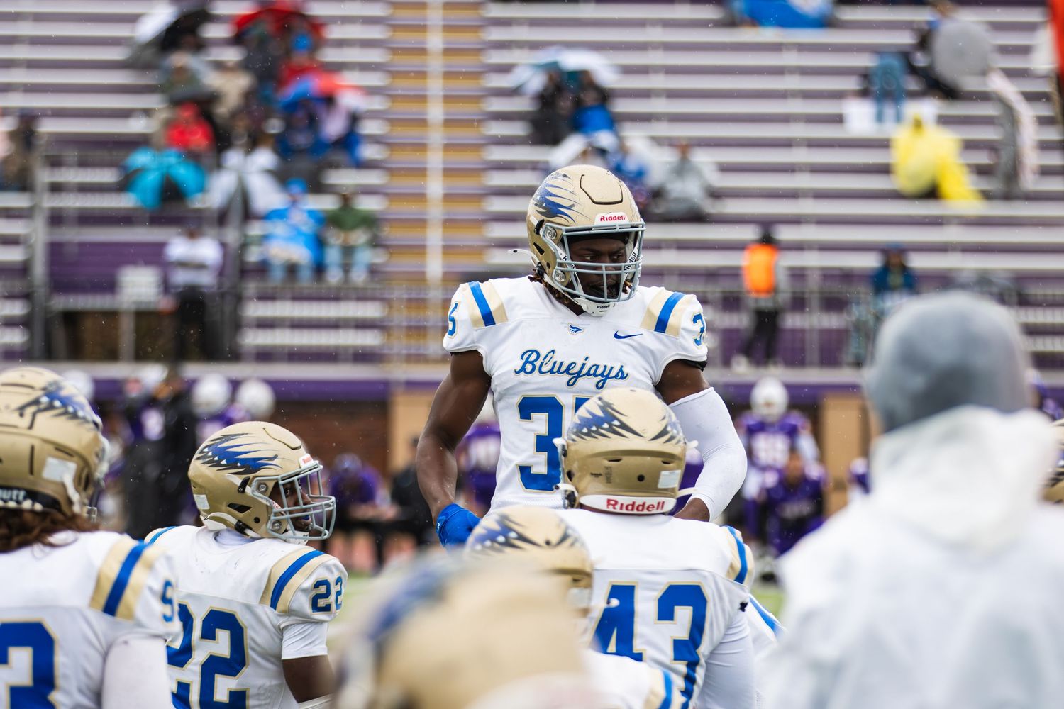 Football players in white and blue uniforms stand on sideline during game.