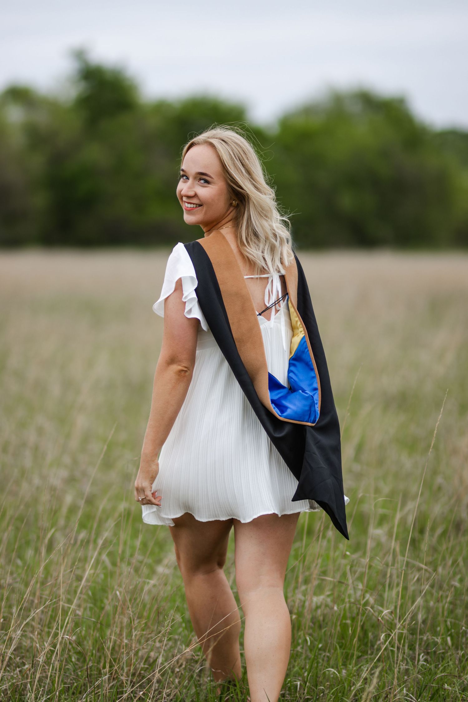 A person in a white dress and dark blazer poses lightheartedly in a tall grass field.