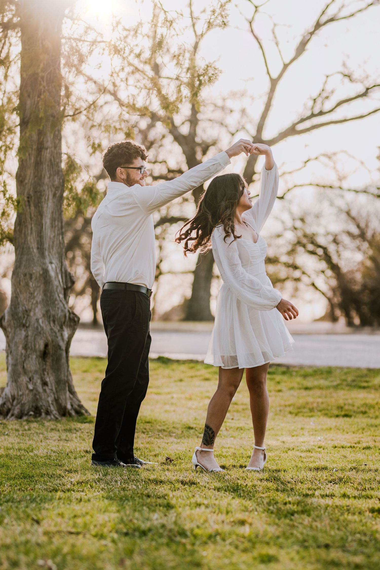 Couple in white dancing together on grass with sunlit trees in background.
