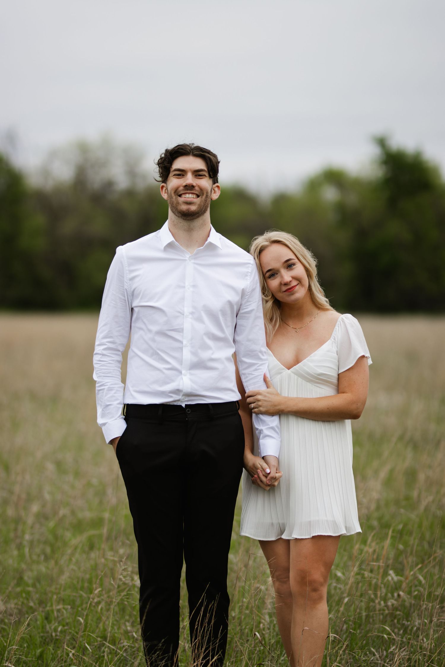 A couple in formal attire poses affectionately in a grassy field with a romantic, natural backdrop.