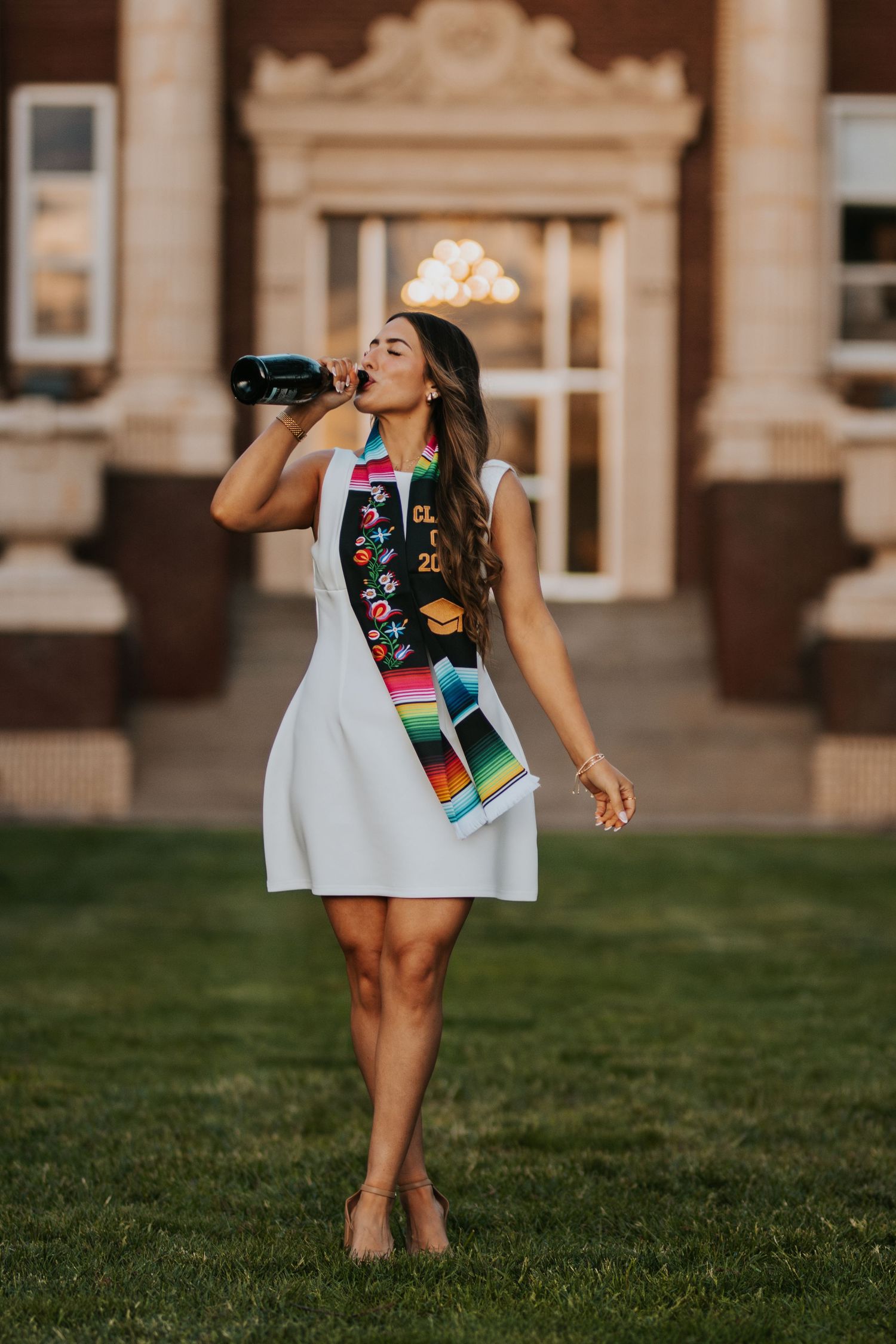 Graduate celebrating with a drink while wearing a decorative sash and white dress on a grassy lawn near ornate architecture.