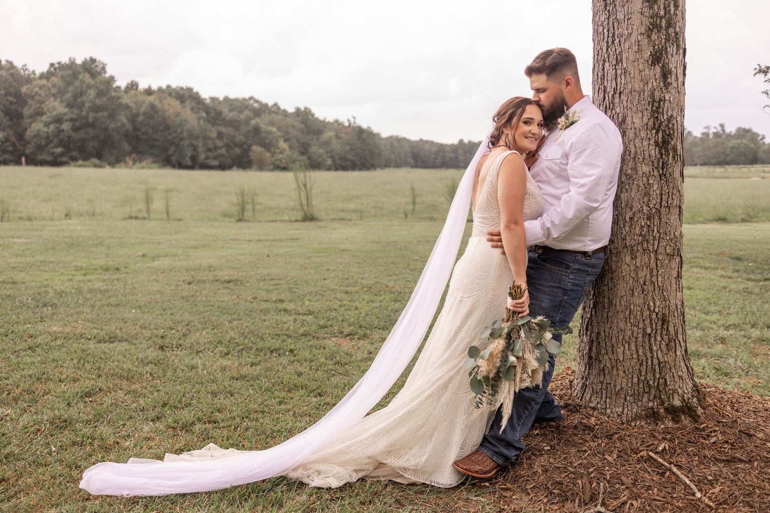 Couple in wedding attire embraces under a large tree in a rustic outdoor setting during a romantic photoshoot.
