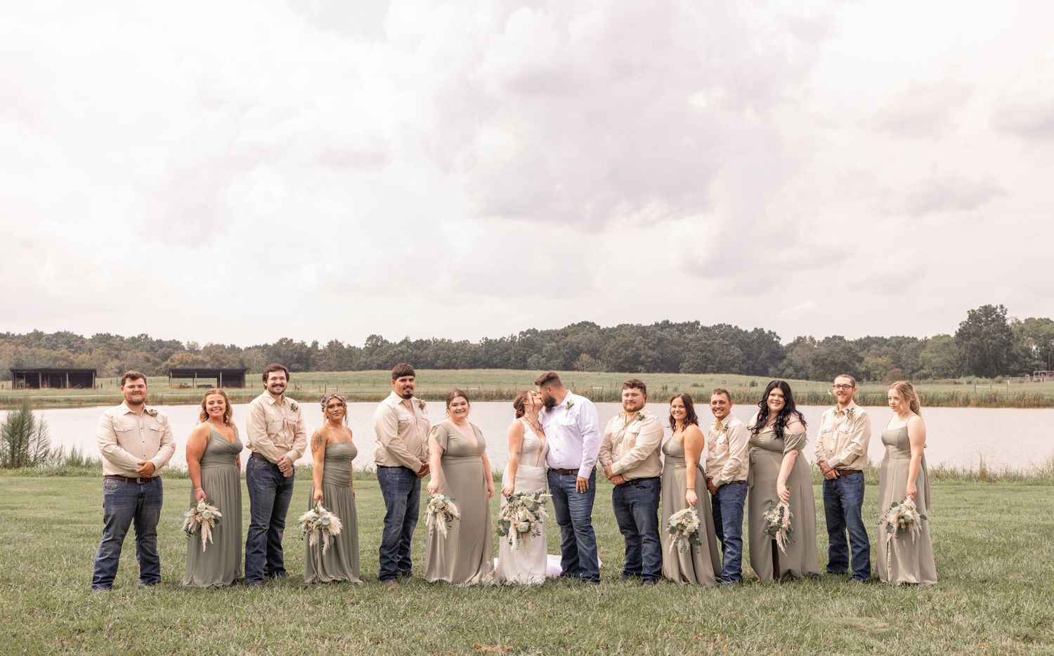 Wedding party in white and beige attire poses together on grassy field near waterfront during outdoor celebration.