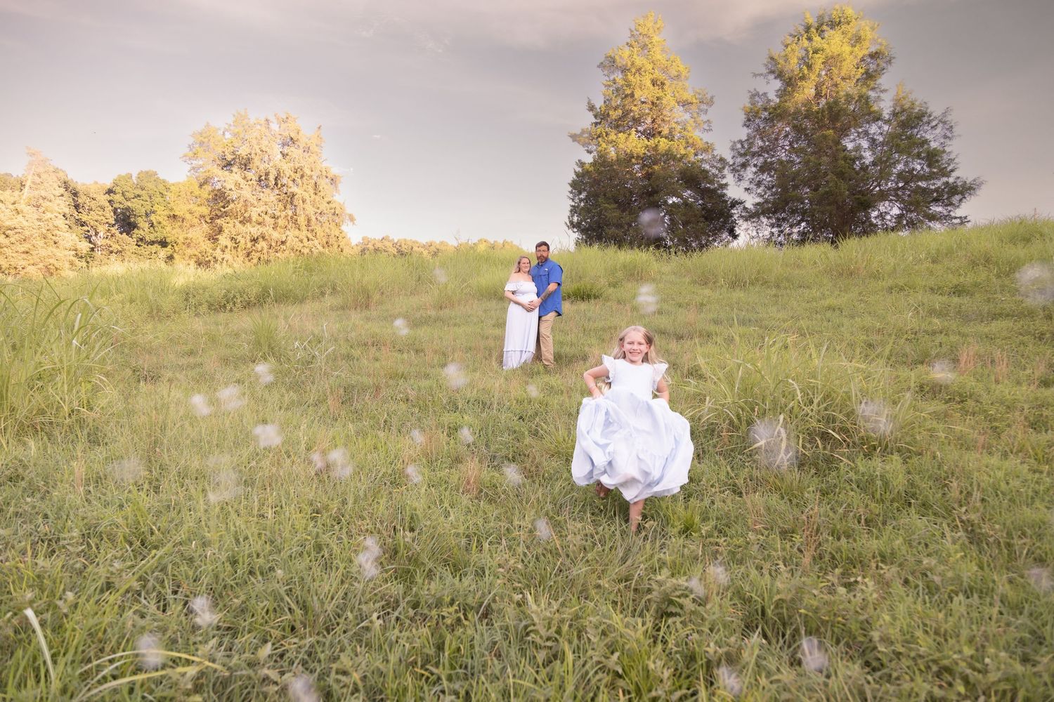 A figure in white clothing walks across a grassy field during golden hour with sunlit trees in the background.