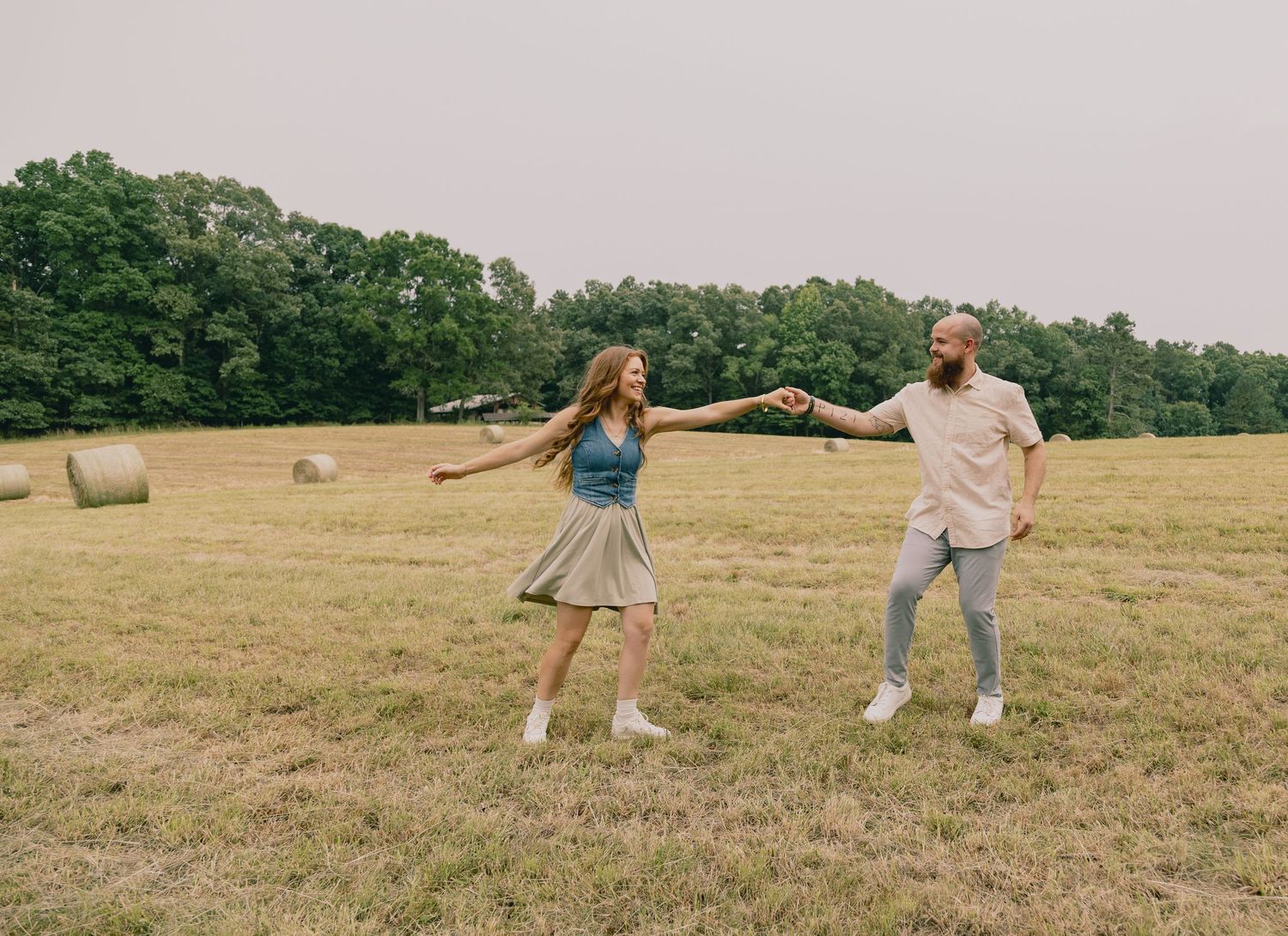 Couple dancing playfully in a field with hay bales on a summer day.