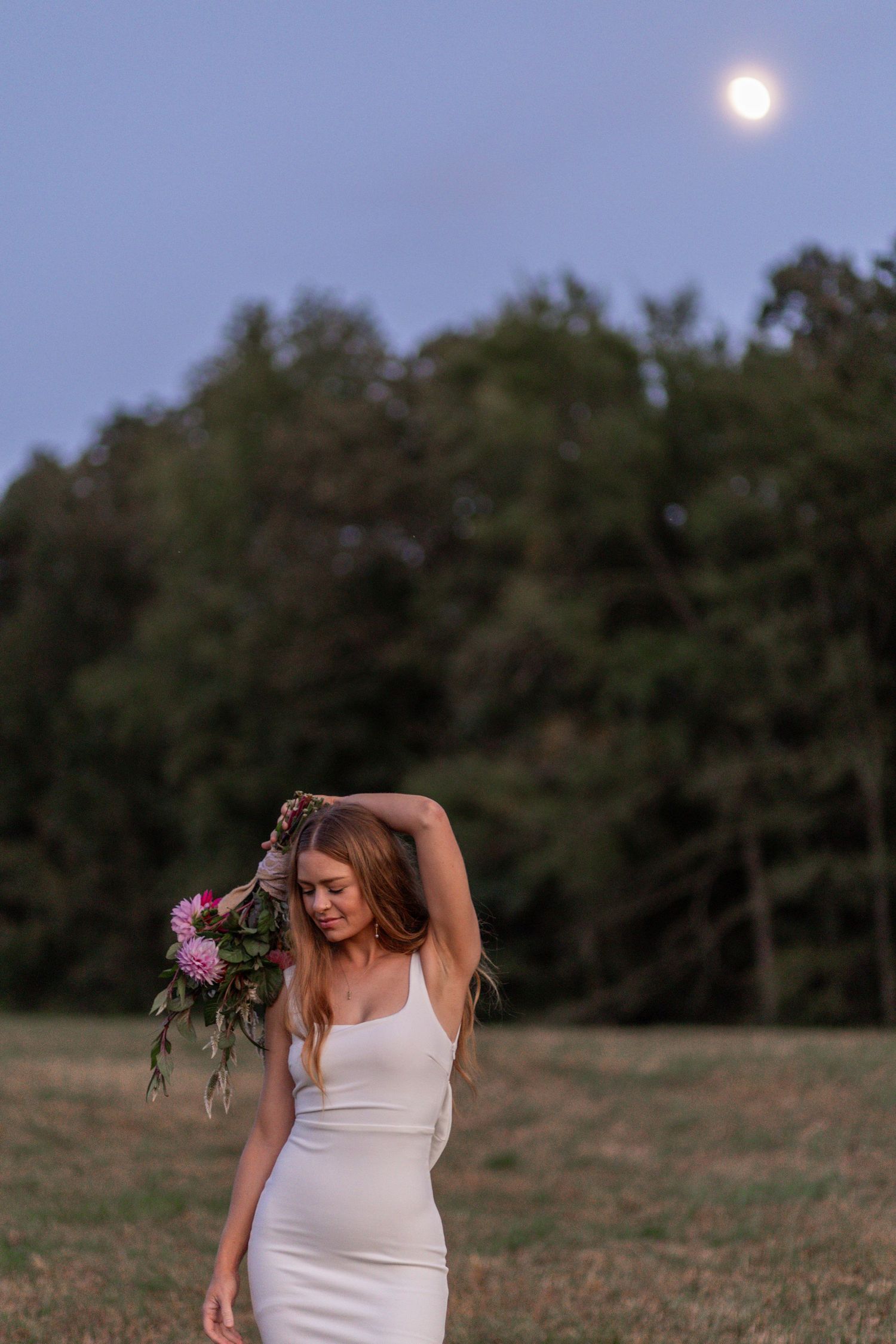 A figure in a white dress stands in a field at dusk with a full moon visible in the darkening sky.