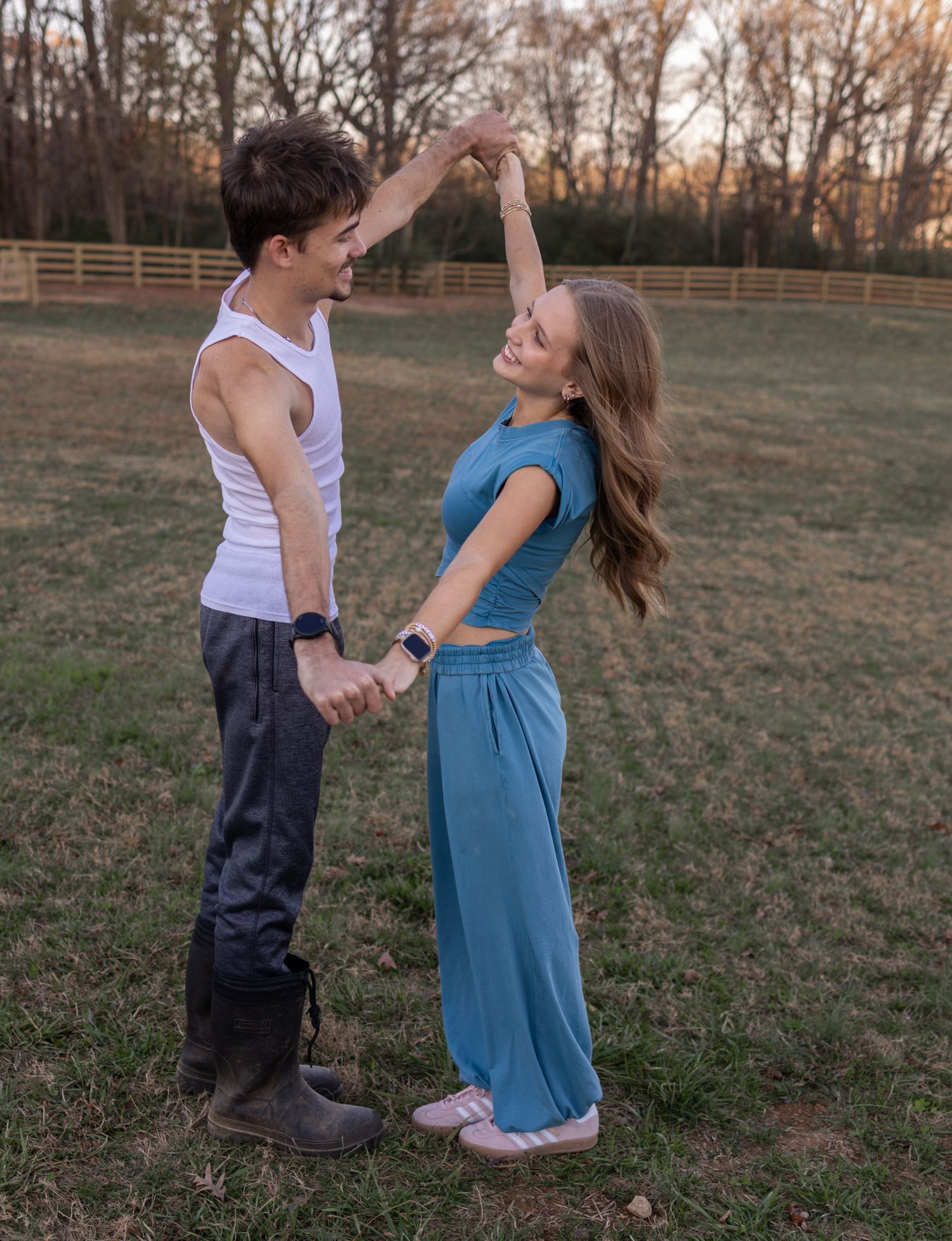 A couple dances together in a grassy field at sunset with a wooden fence in the background.