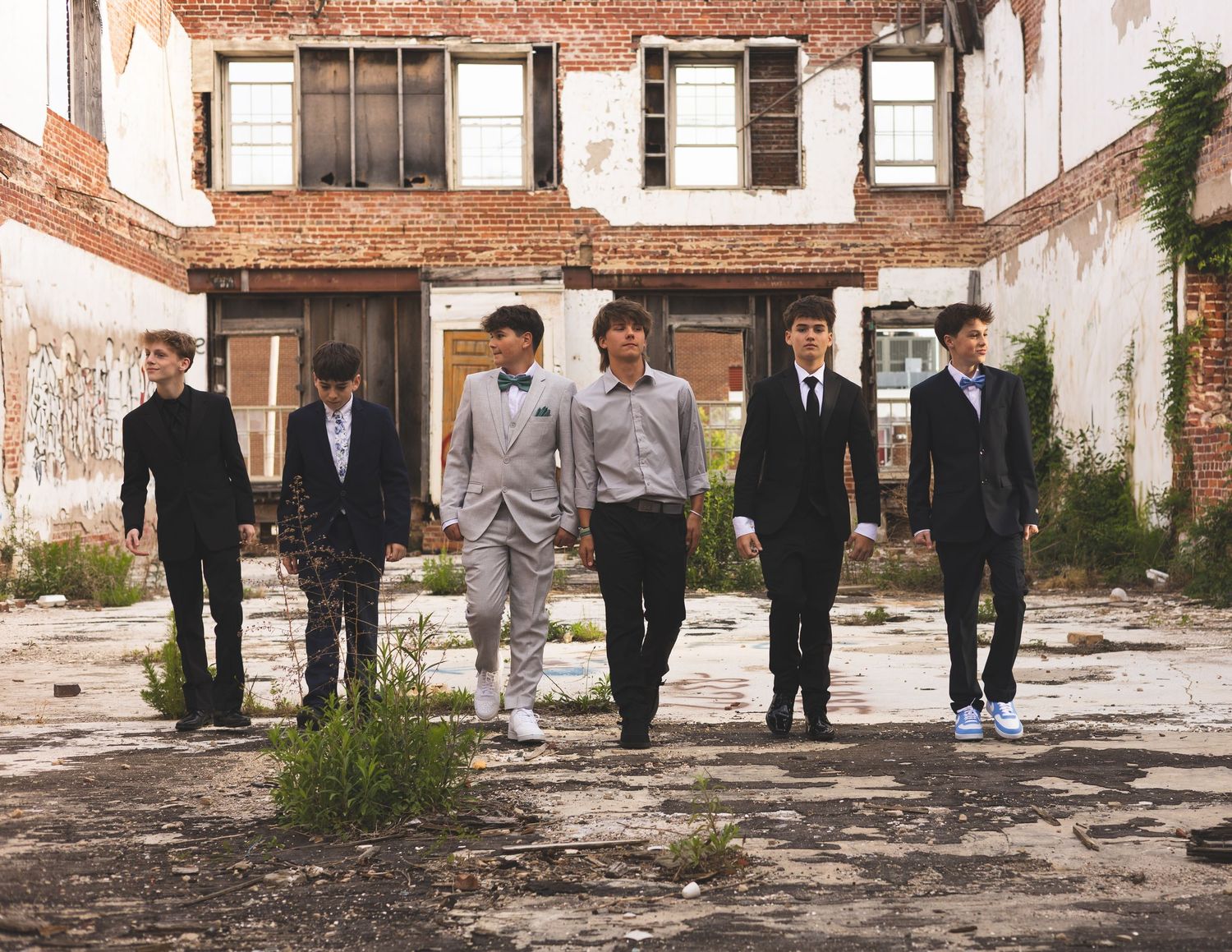 Six young men in formal suits pose in an abandoned urban courtyard with weathered brick building backdrop.