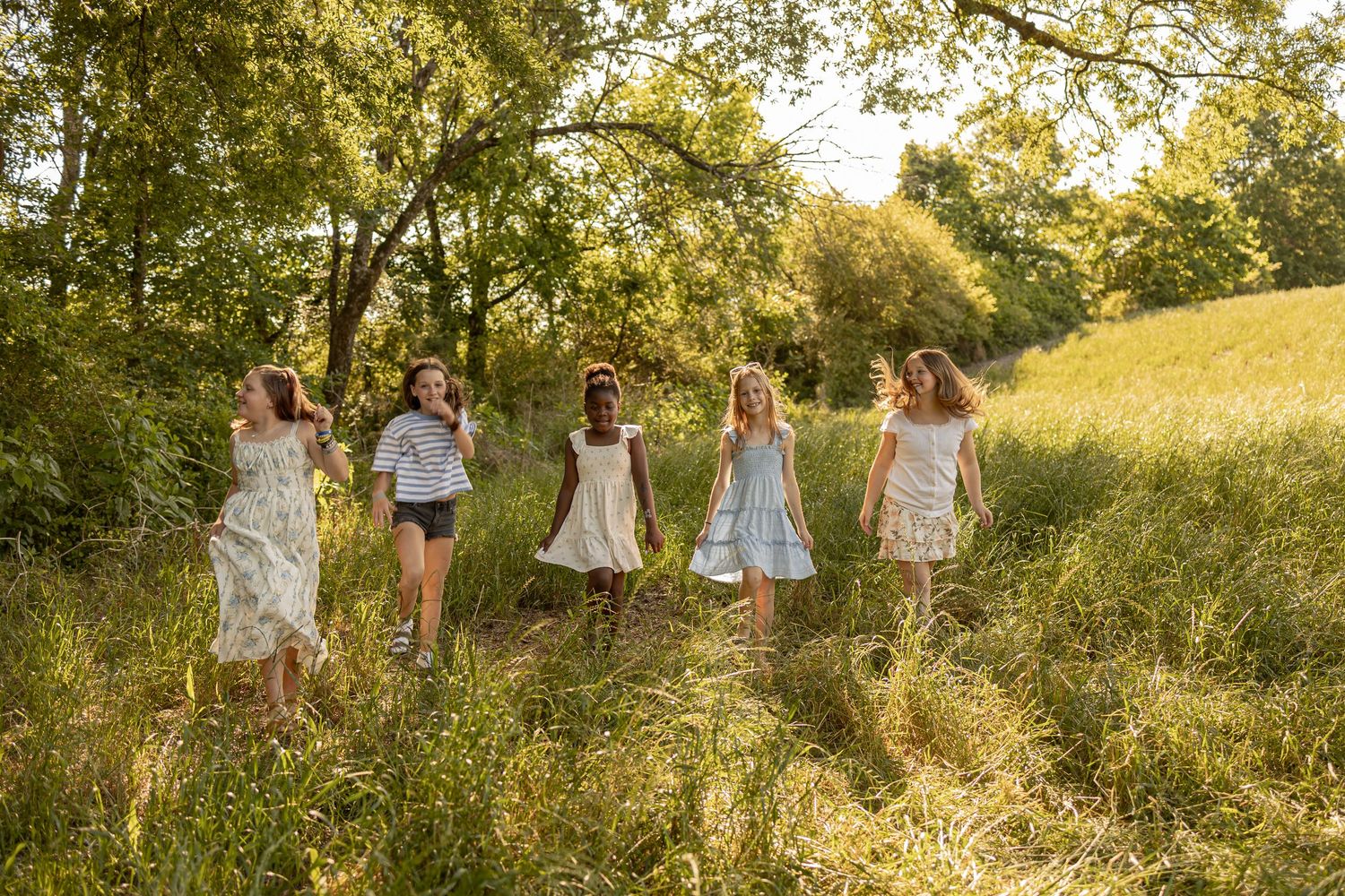 Six girls in summer dresses walk together through a sunlit meadow path surrounded by lush green trees and tall grass.