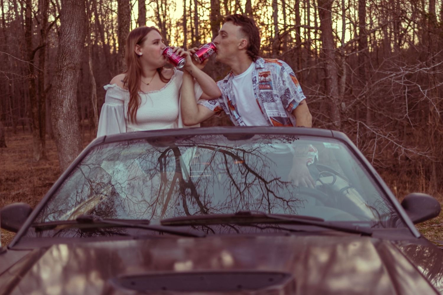 A young couple stands through a car sunroof in a forest, drinking from cans at sunset.