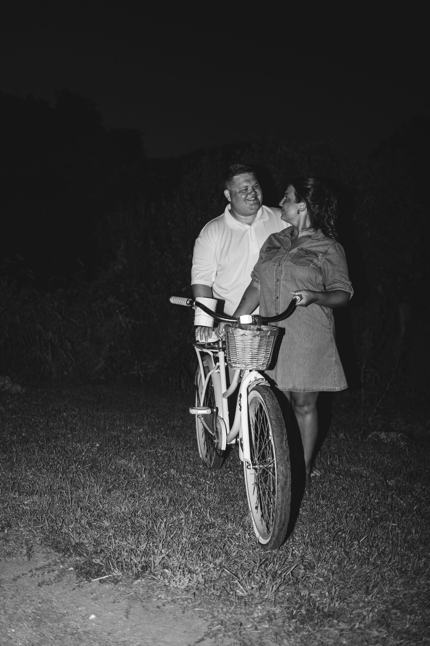 Black and white photo of a romantic couple standing together with a bicycle at night outdoors.