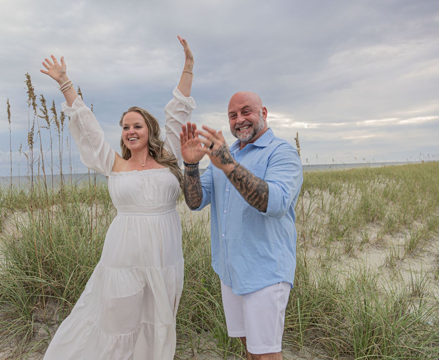 Joyful couple celebrating on a beach with sea grass and cloudy sky in the background.