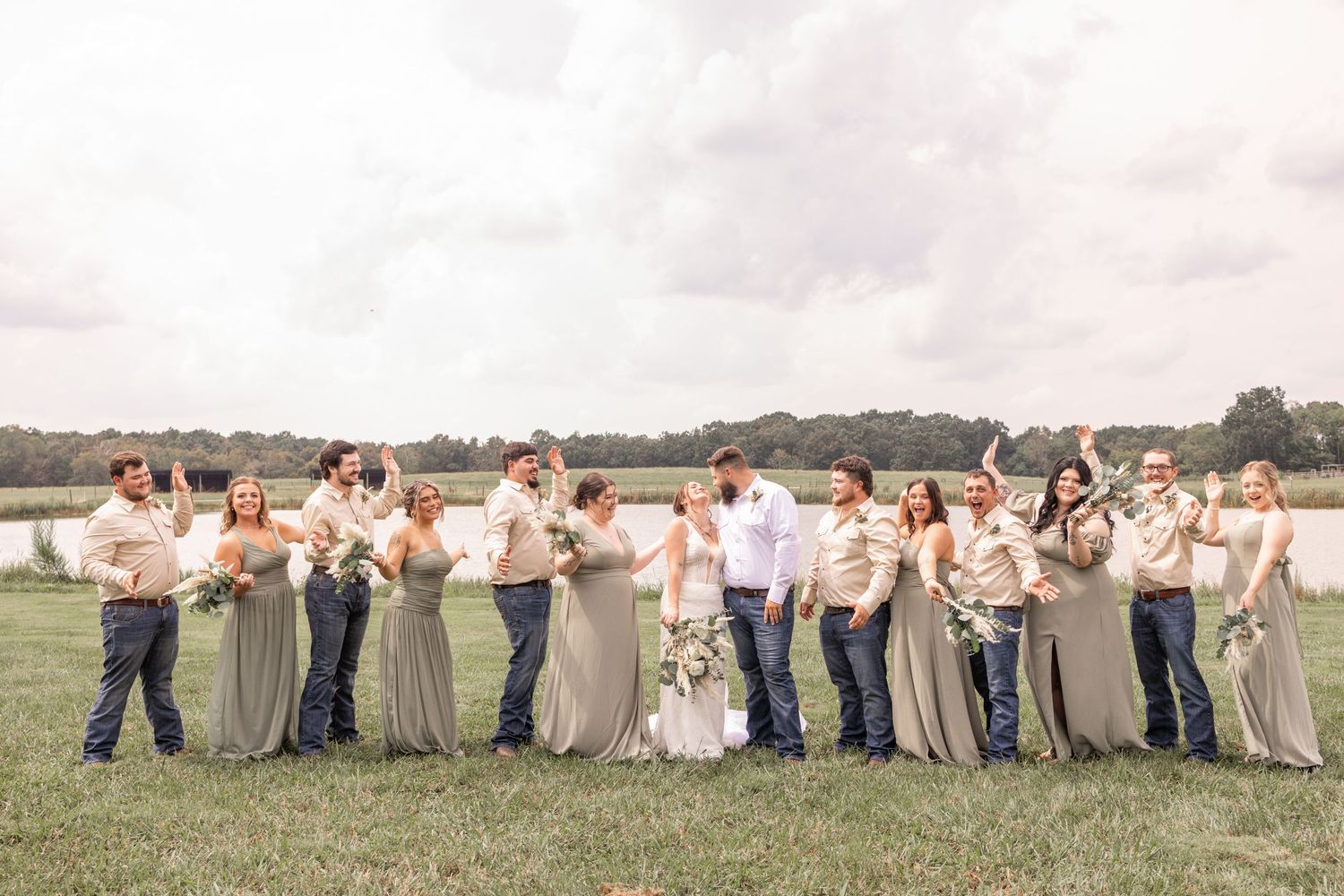 Wedding party in beige and white outfits jumps joyfully in unison on a grassy field at sunset.