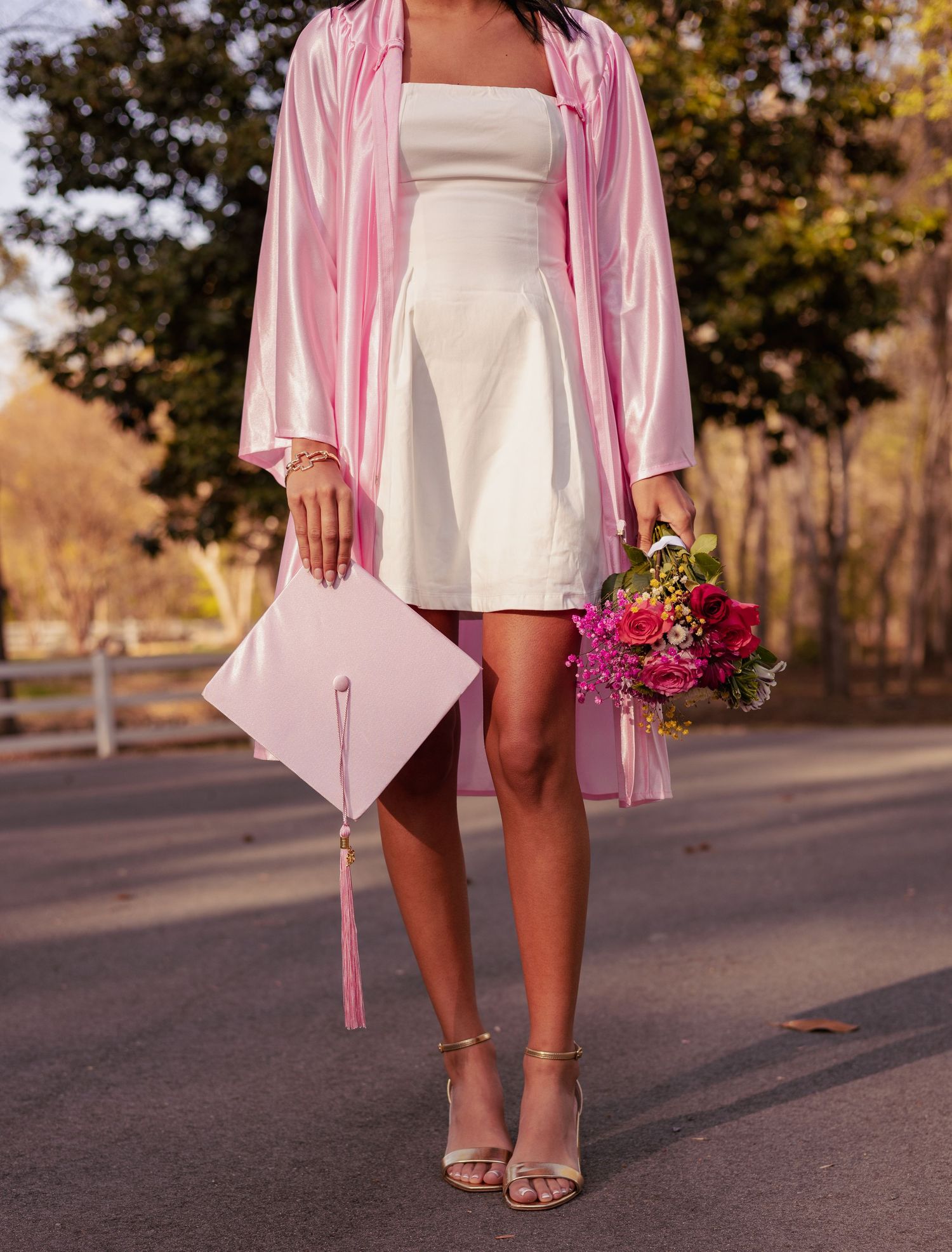 A graduate in a pink cap and gown holds a bouquet of pink flowers while wearing a white dress and gold heels.