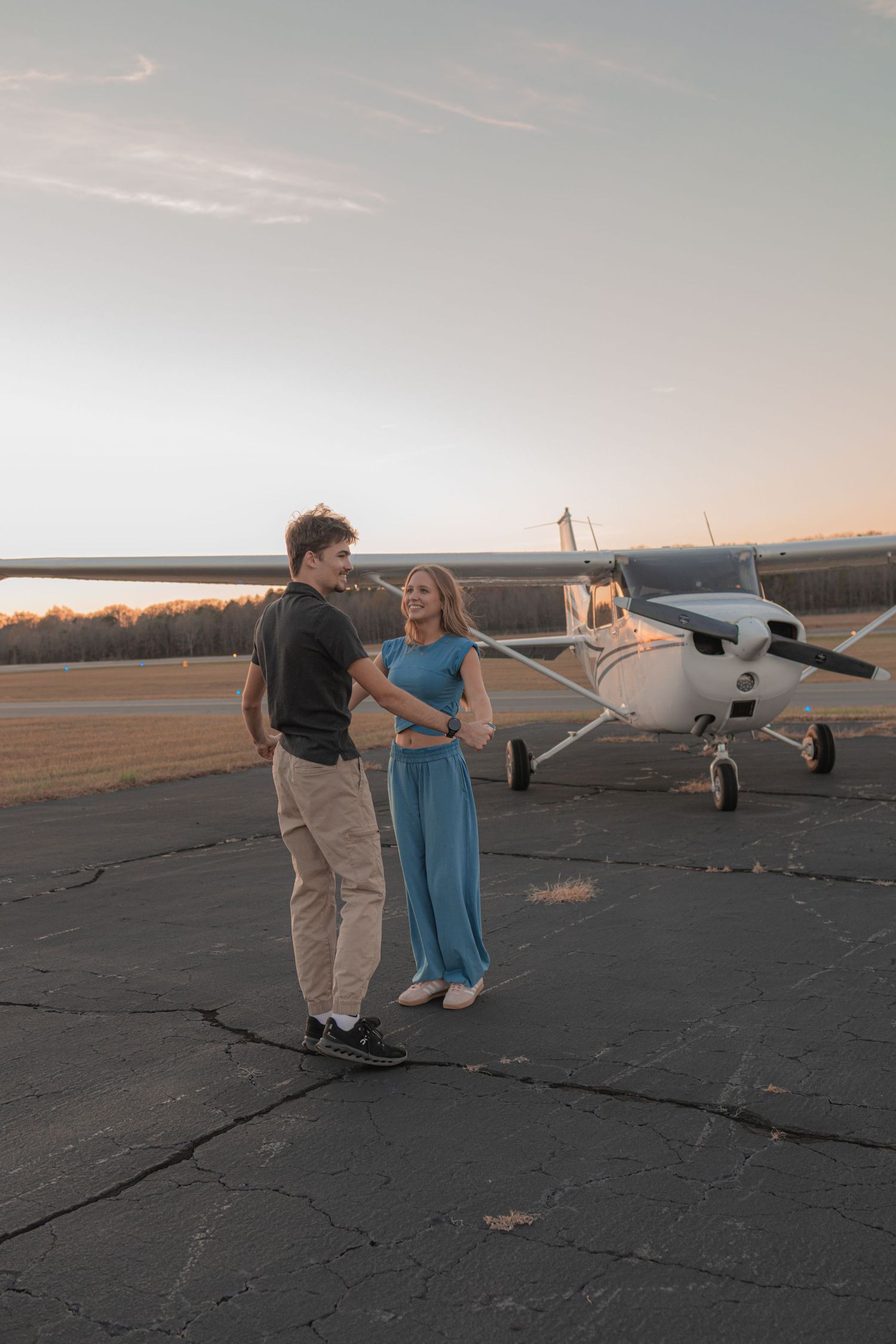 A couple stands together on an airfield at sunset with a small private aircraft in the background.