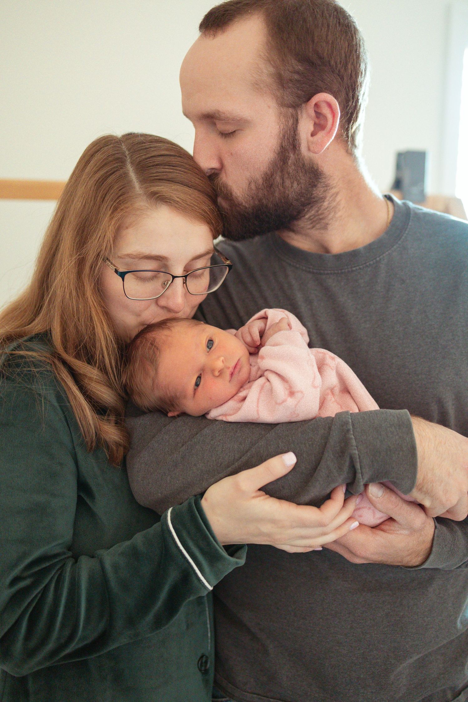 A tender family moment as parents lovingly embrace their newborn baby wearing a pink onesie.