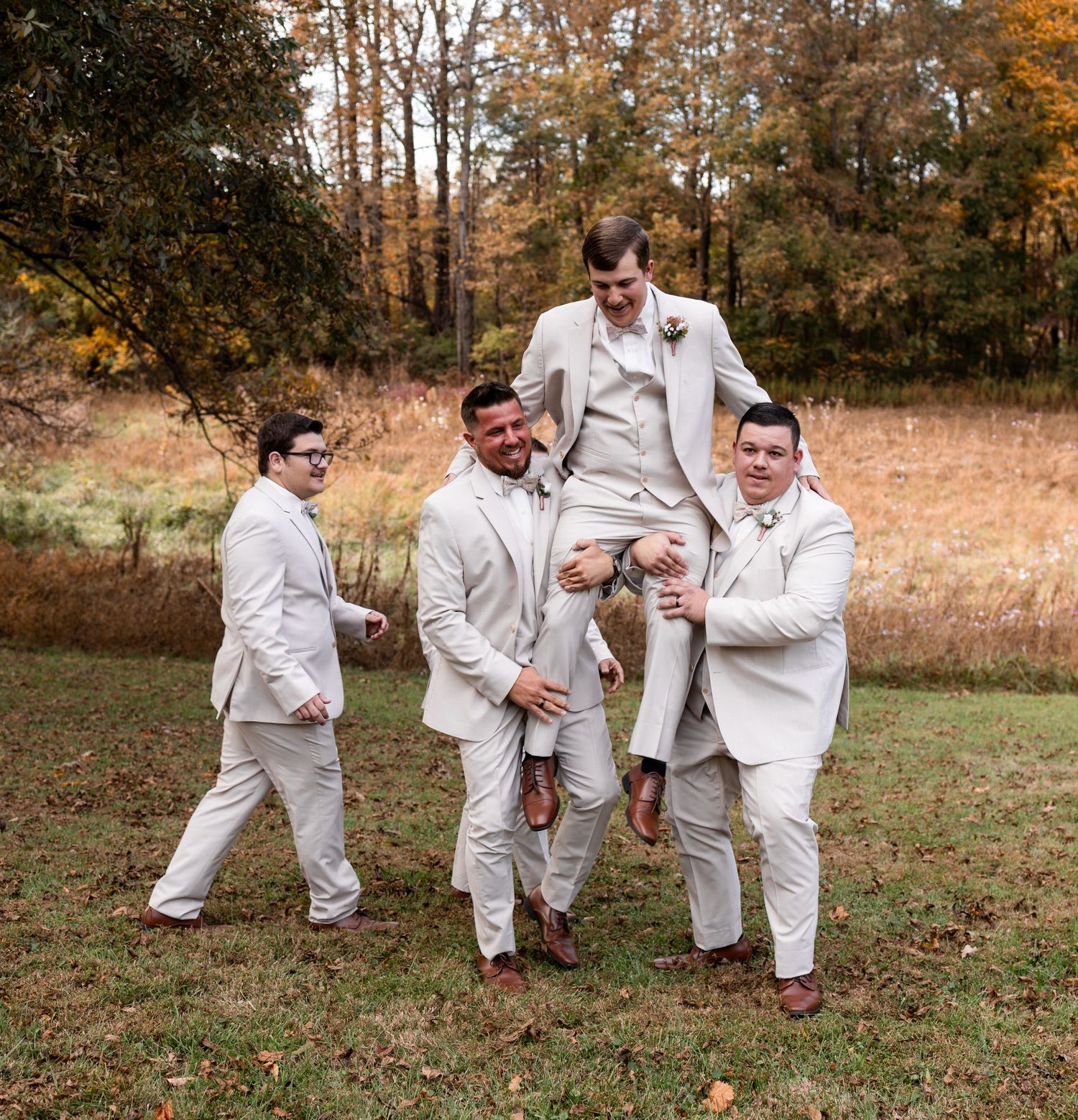 Groomsmen in cream suits lifting the groom outdoors during a fall wedding celebration.