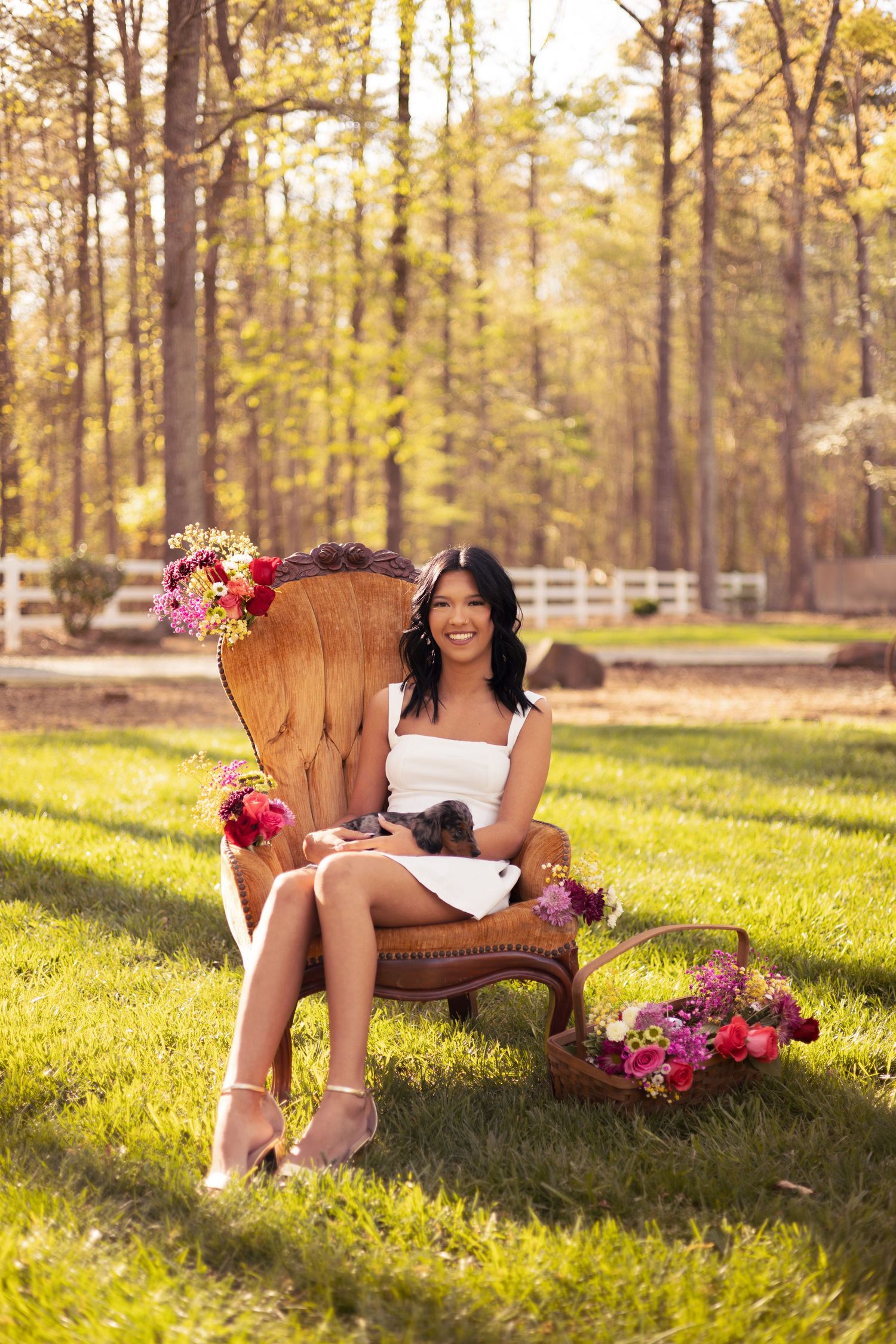 Woman in white dress sitting in ornate chair surrounded by colorful flowers on lush green lawn with trees in background.