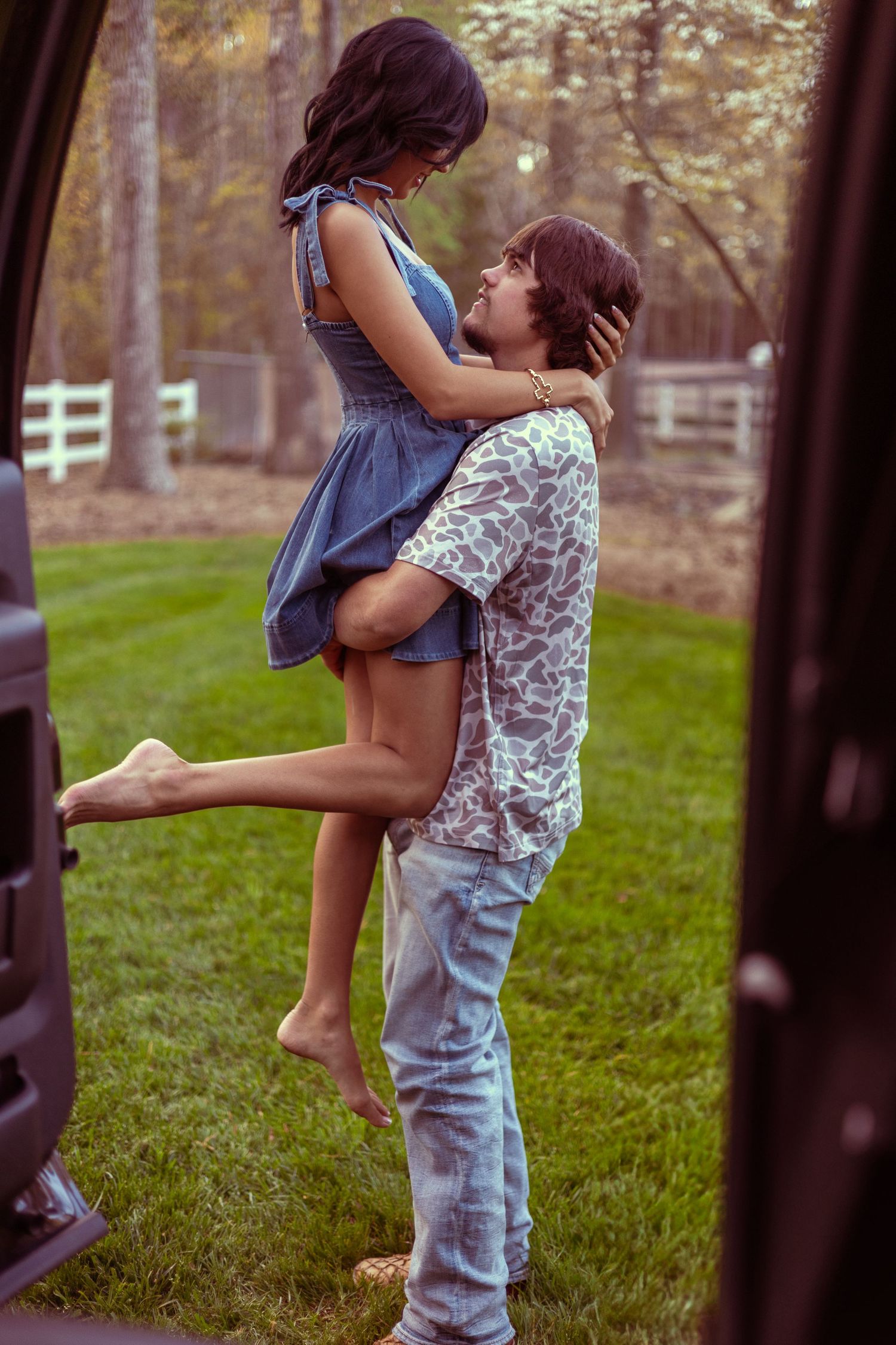 A man lifts a woman in a blue dress outdoors near a vehicle, sharing a romantic moment on a green lawn.