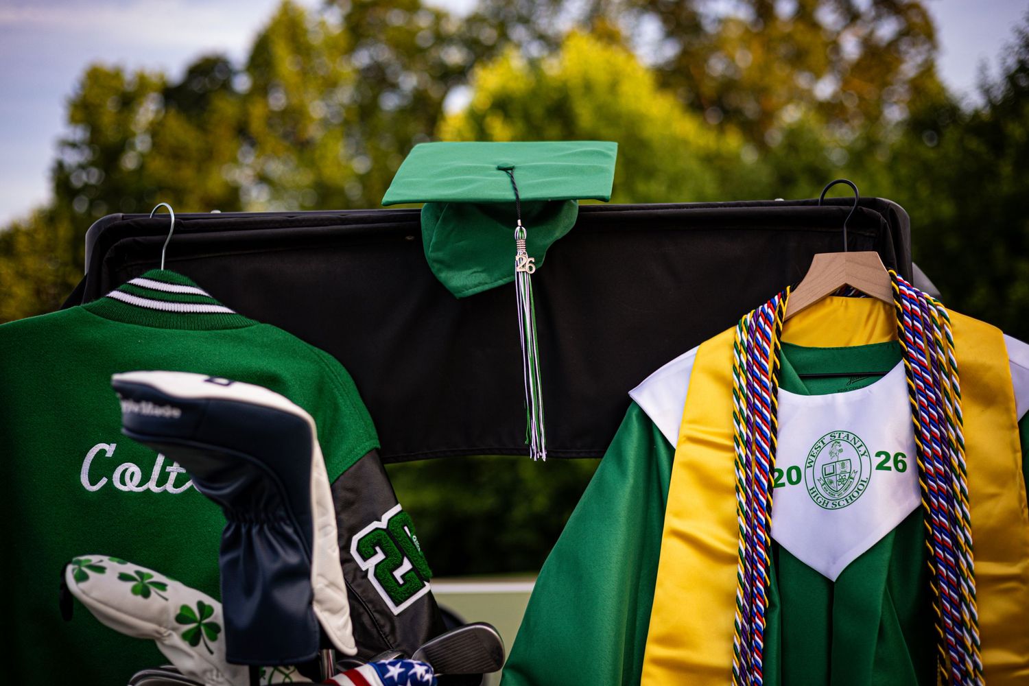 Green graduation cap, gown with honor cords, and varsity jacket displayed on hangers outdoors for class of 2026.
