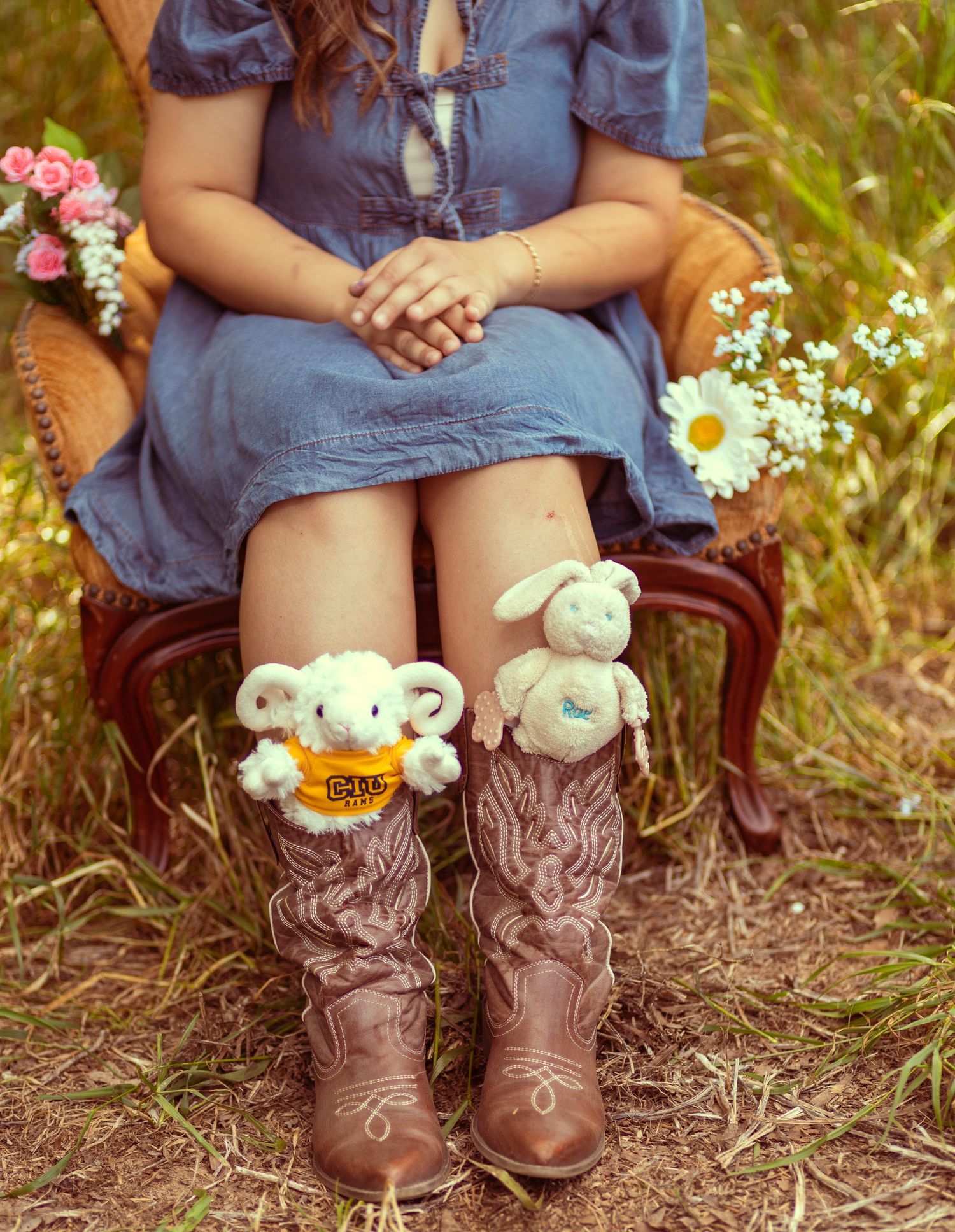 Woman in blue dress sitting in chair wearing cowboy boots adorned with cute plush lamb stuffed animals outdoors.