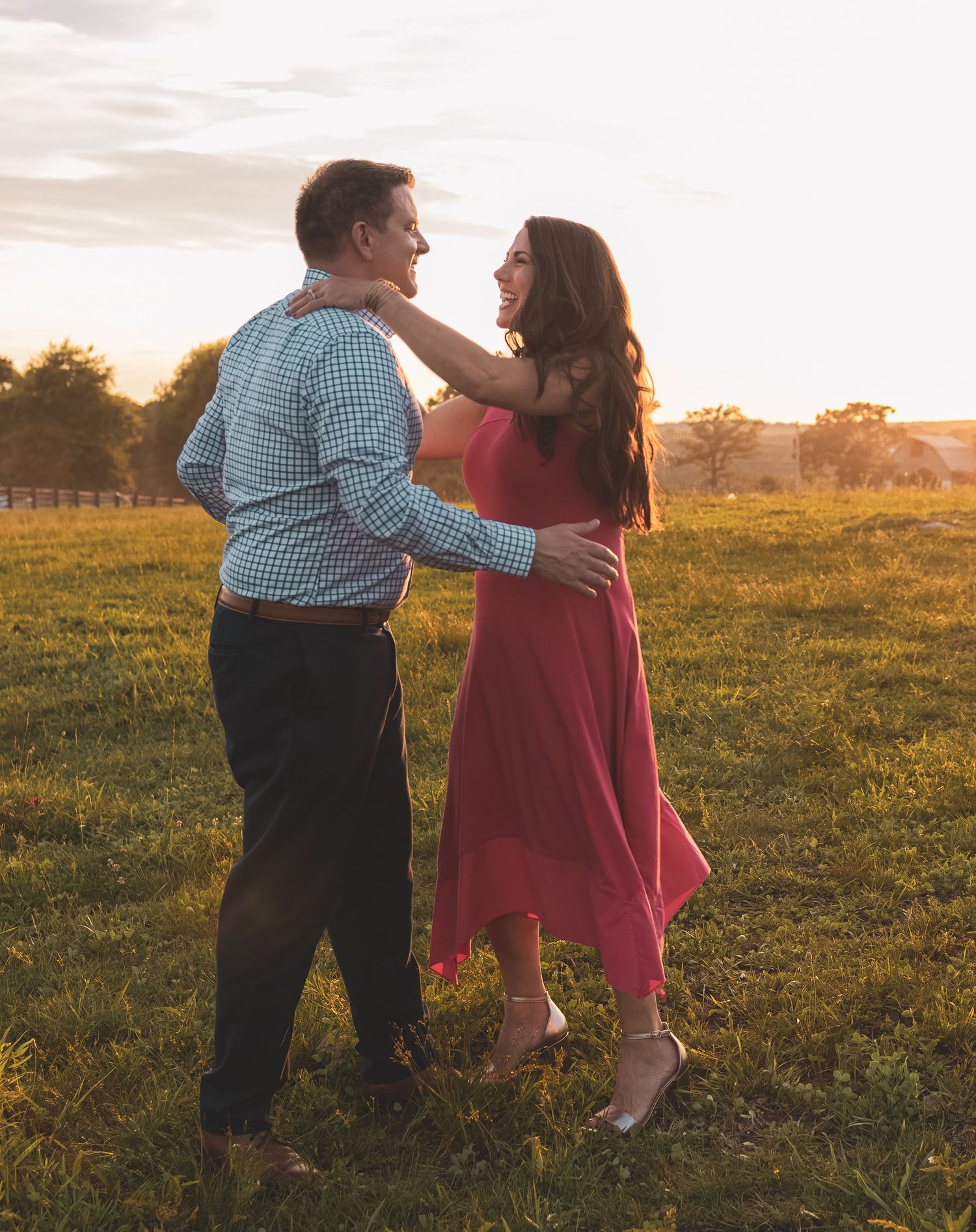 Couple dancing together in a green field at golden hour sunset, woman in red dress laughing joyfully.