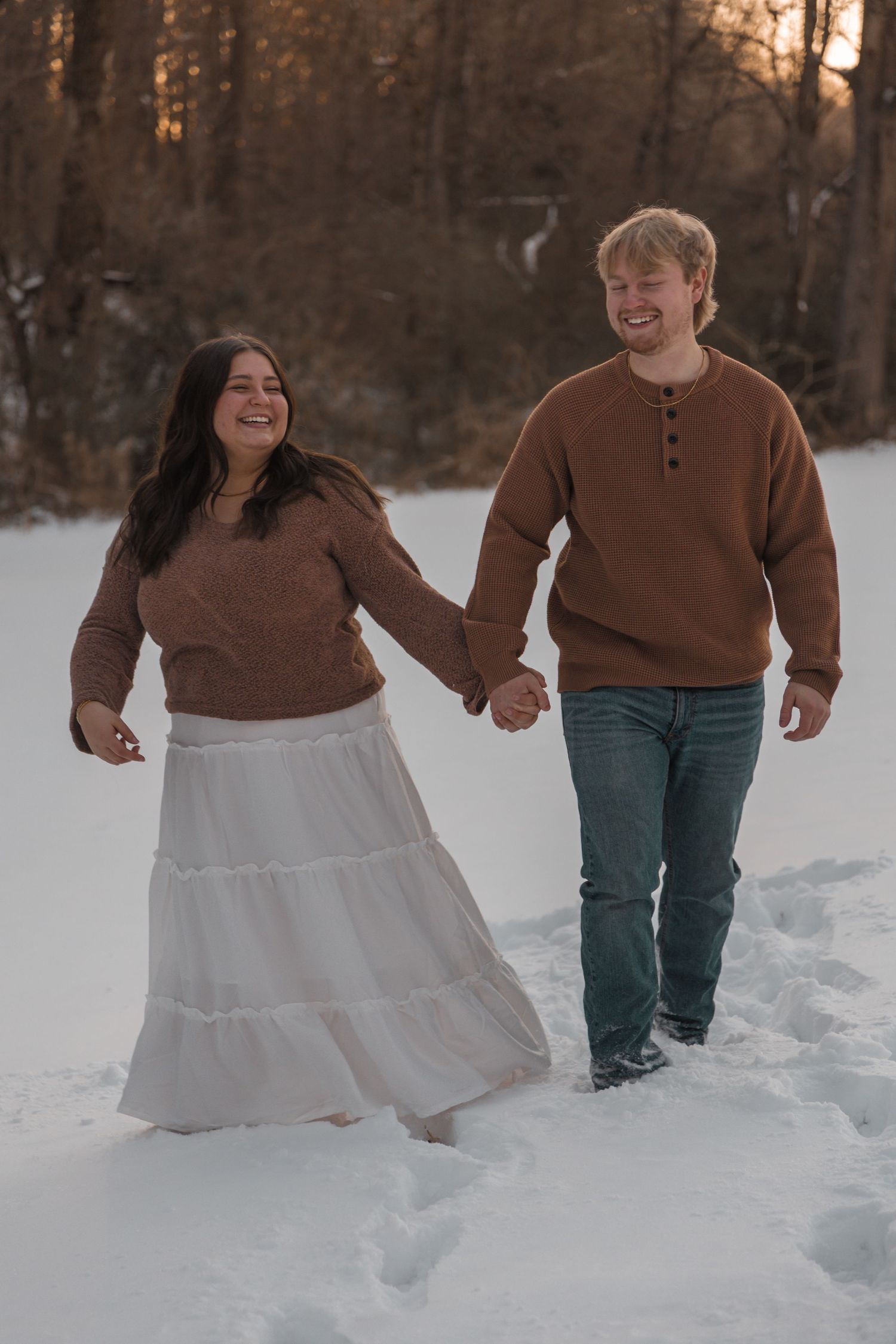 Couple holding hands walking in snow wearing matching brown sweaters, white skirt, and jeans with winter trees at sunset.