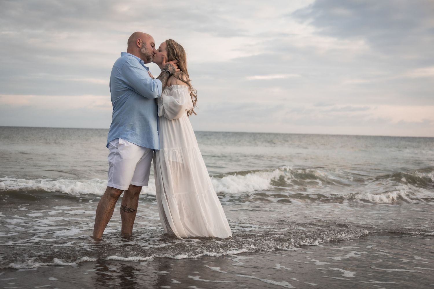 A romantic couple shares a tender kiss while standing in the ocean waves at sunset, wearing elegant beach attire.