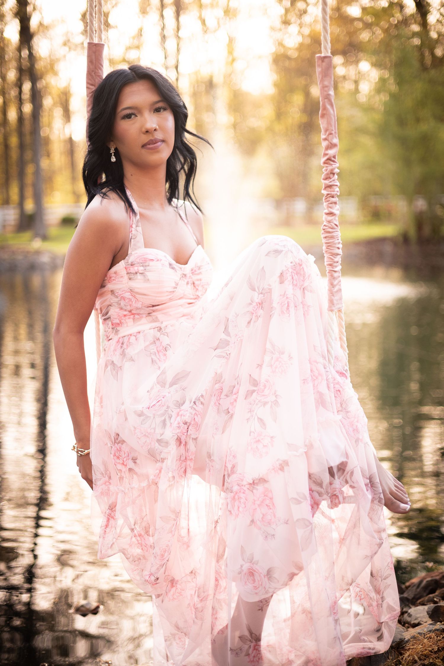 Woman in pink floral gown standing on swing by reflective pond in golden sunlight.
