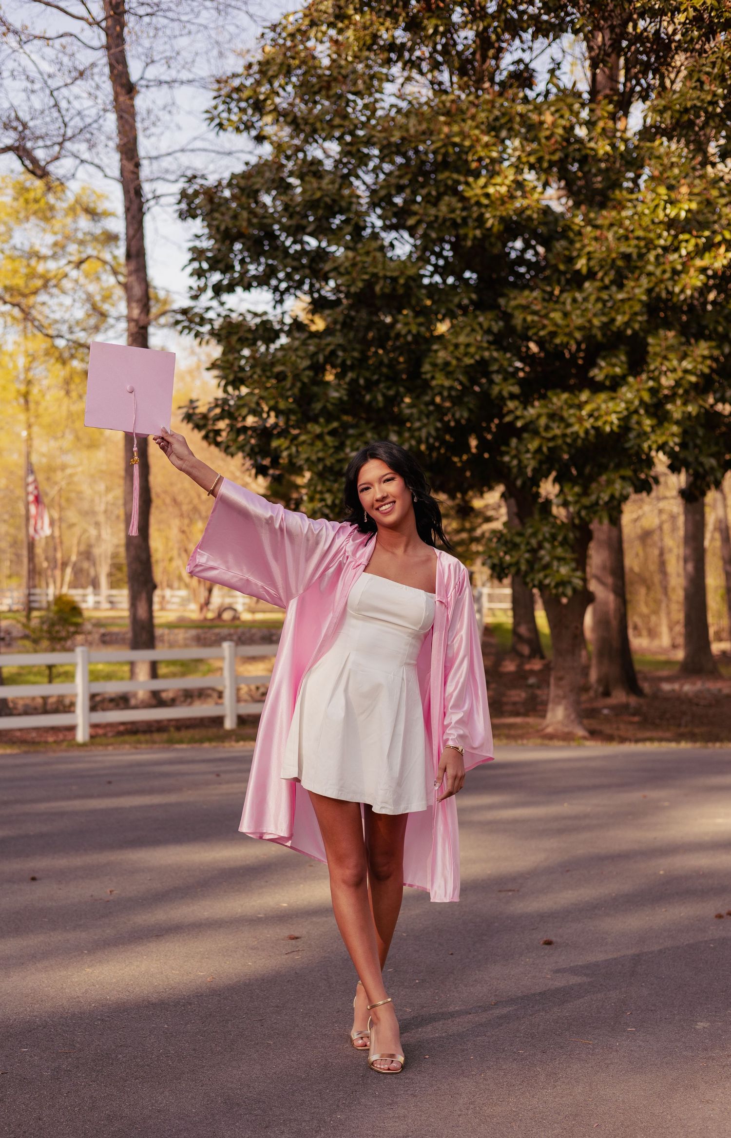 Young woman in white dress and pink graduation gown holds cap up celebrating outdoors among trees.