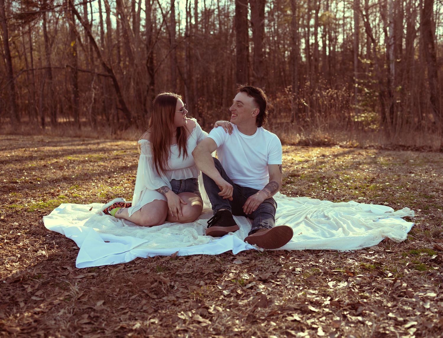 Young couple sitting on white blanket in autumn forest, enjoying romantic outdoor picnic together.
