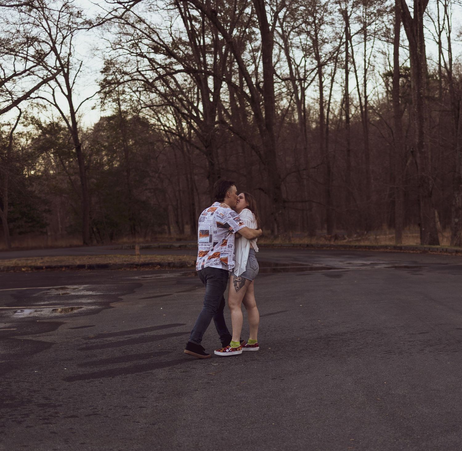 A couple embracing and walking together in an empty parking lot surrounded by bare trees in a park.