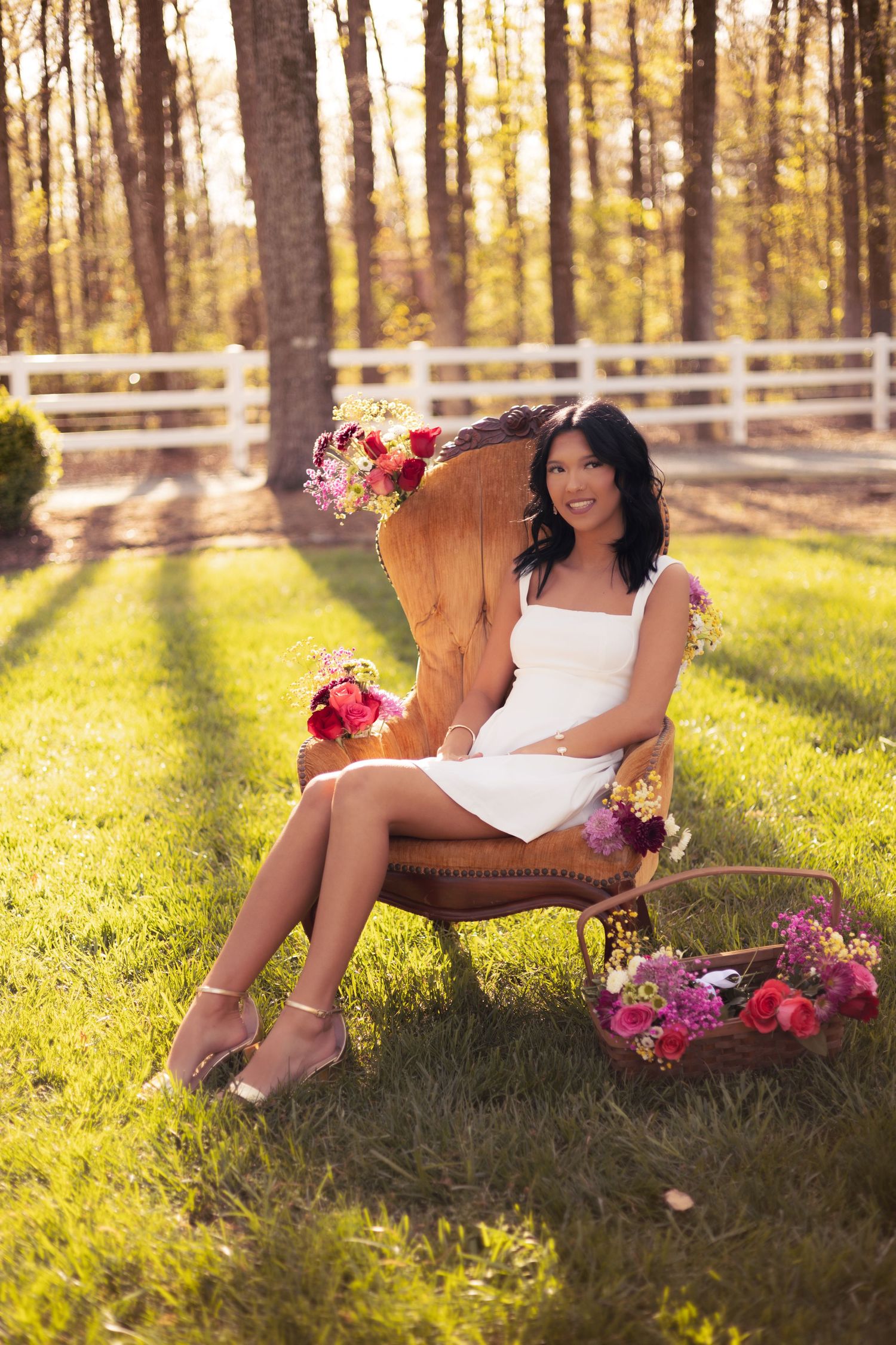 Woman in white dress sitting in vintage chair outdoors surrounded by colorful flowers with white fence and trees in background.
