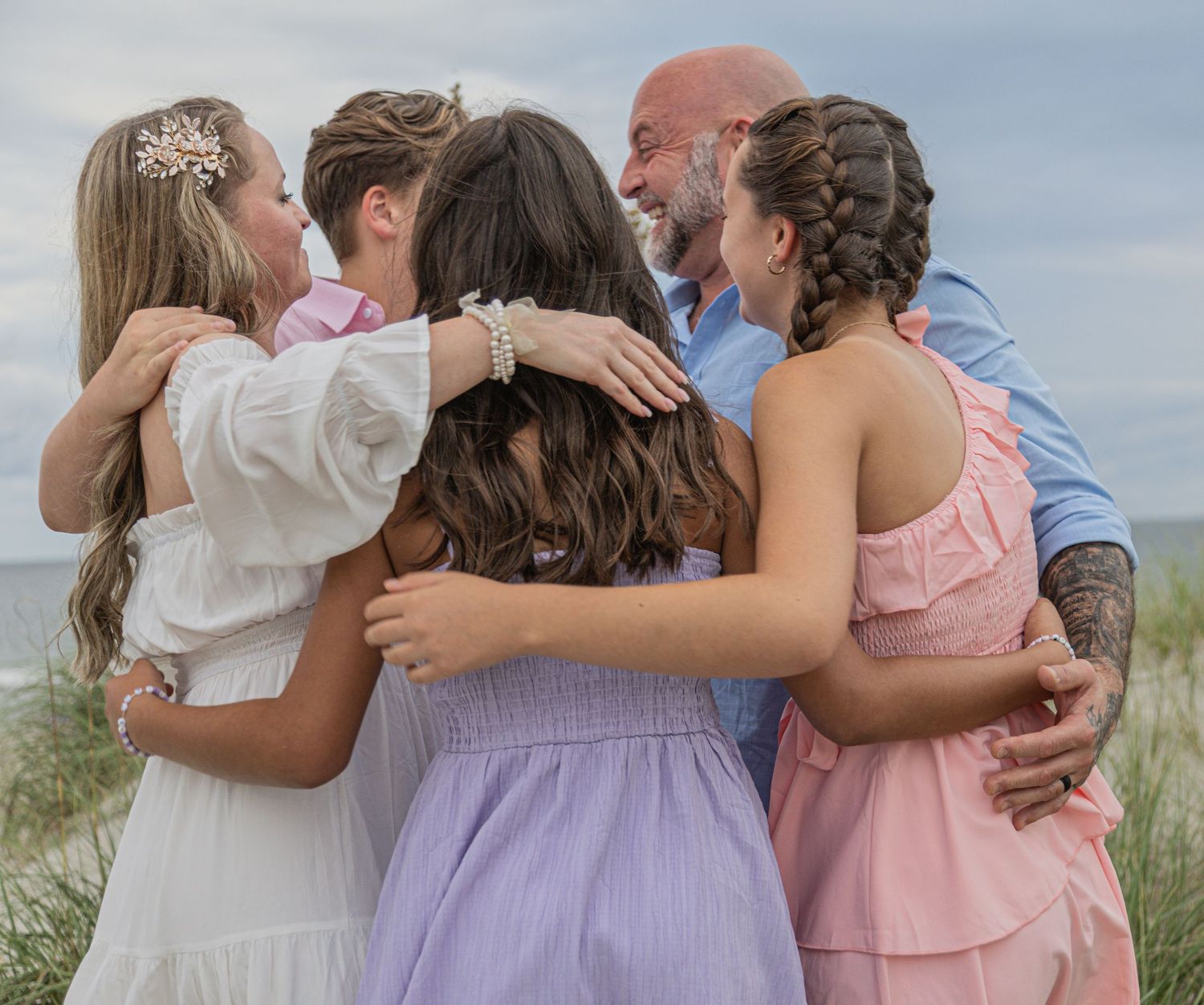 Group in pastel dresses embracing in a heartfelt moment on sandy dunes under cloudy skies.