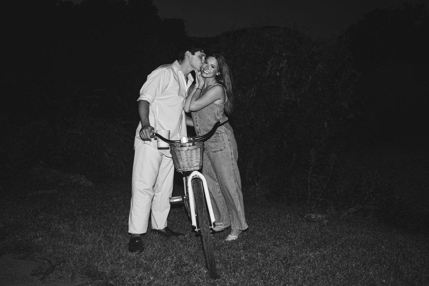 Couple sharing a romantic kiss outdoors at night beside a bicycle with basket, black and white photo.
