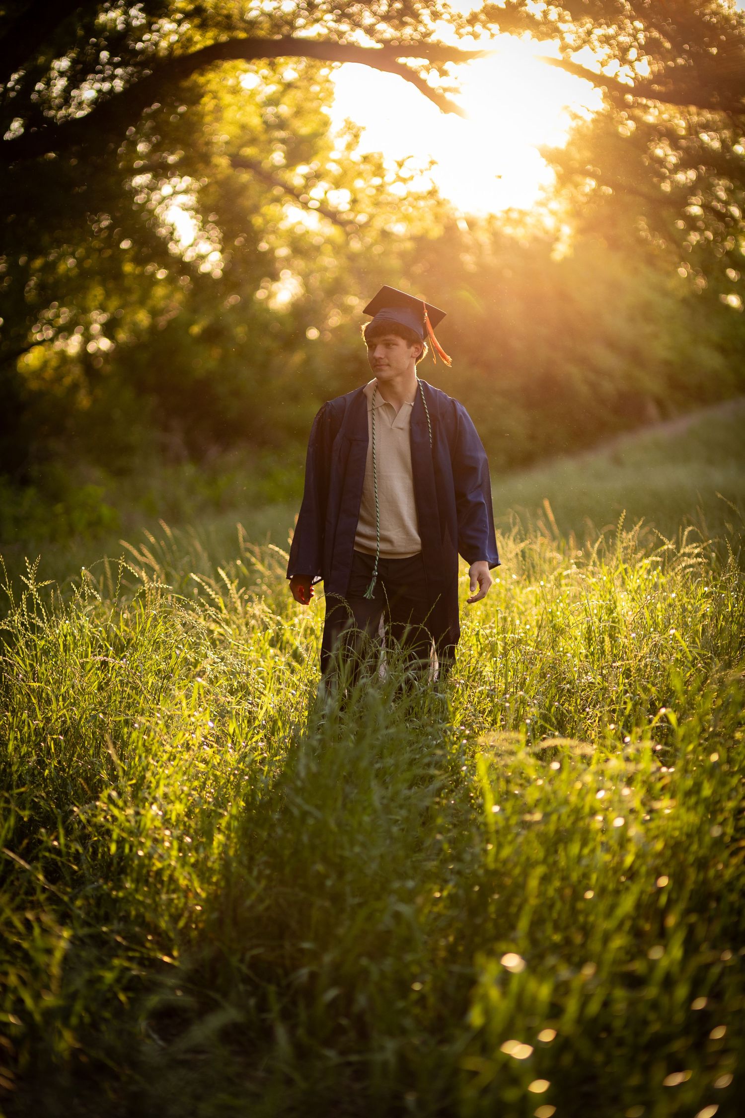 Graduate in cap and gown walking through lush green field at golden sunset.