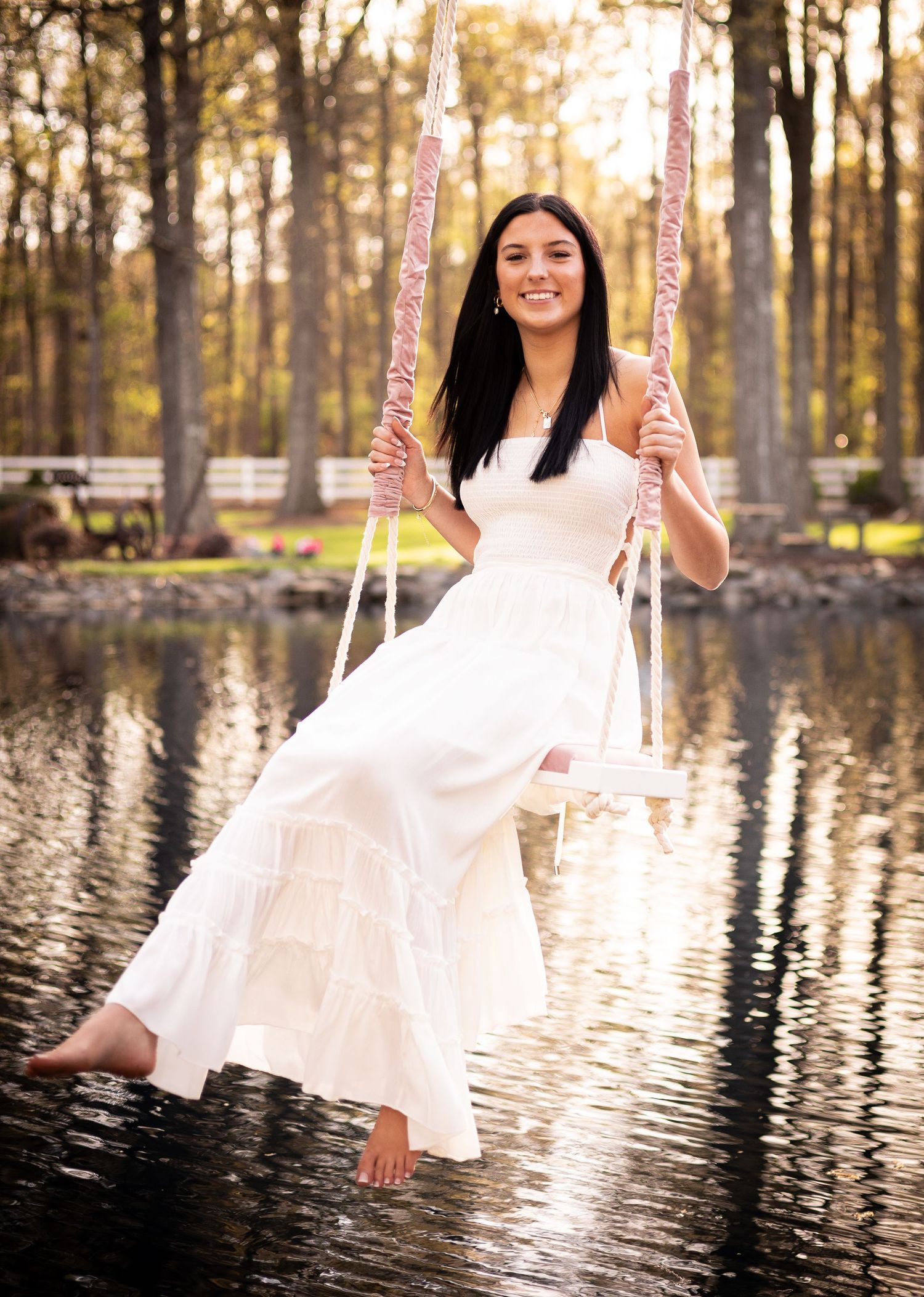 Young woman in white dress sitting on a swing over a reflective pond surrounded by trees.