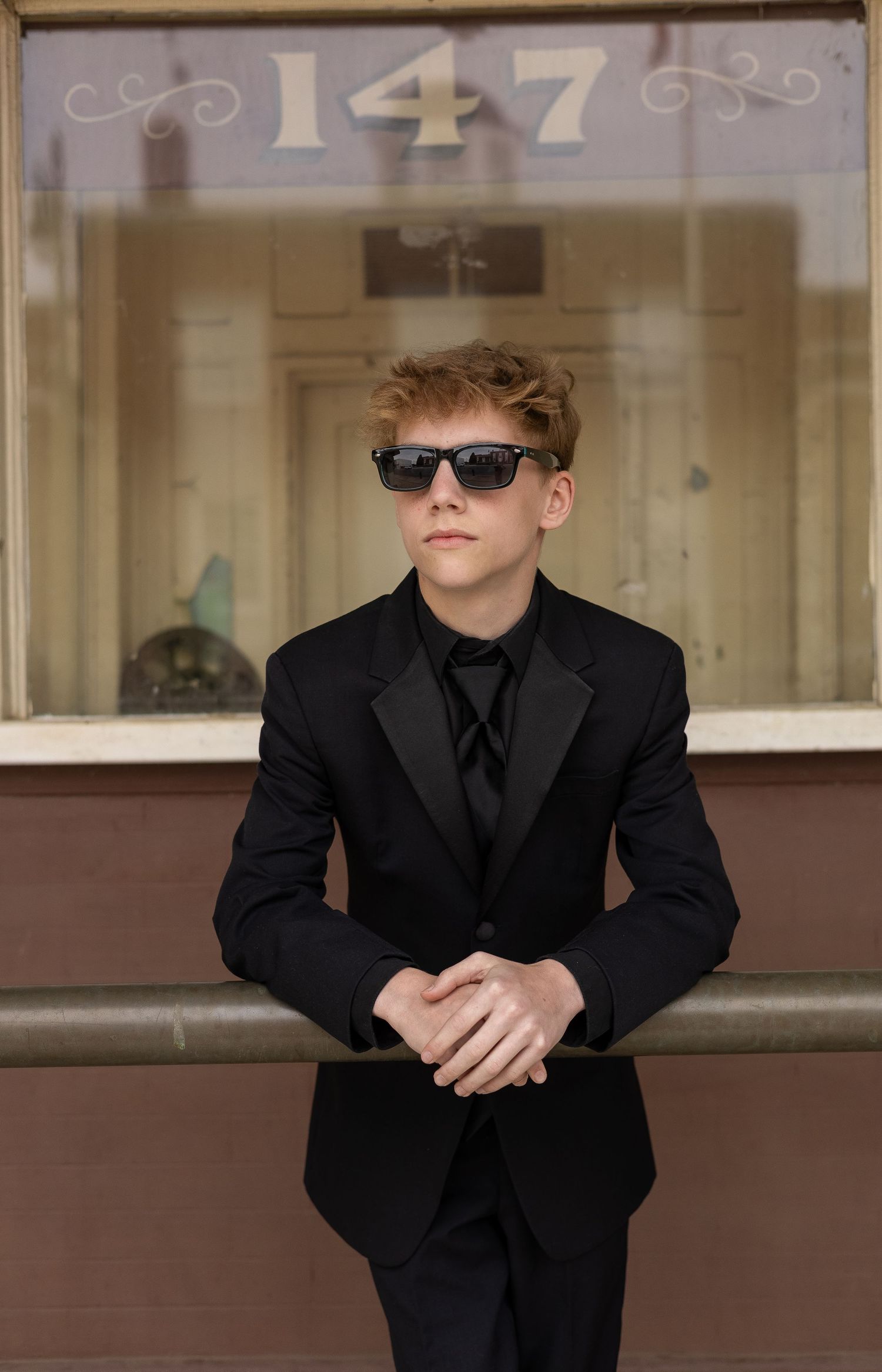 Young man in black suit and sunglasses leaning on railing outside building numbered 147.