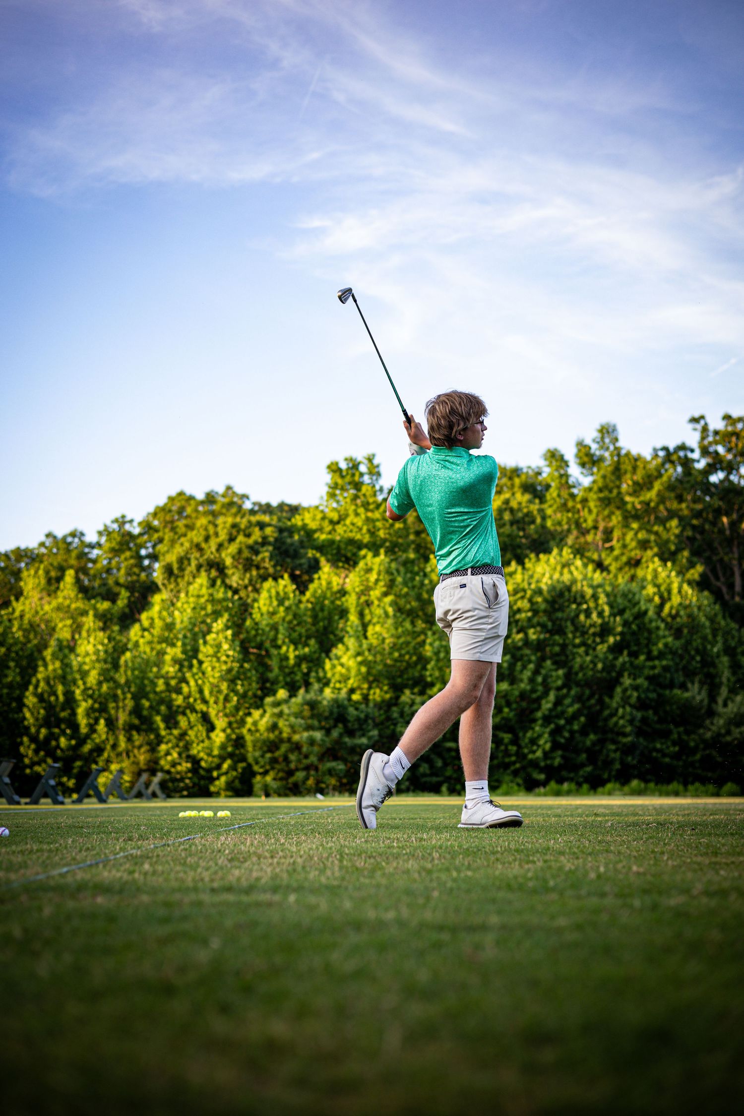 Young golfer in green polo and white shorts completing a swing on a lush golf course with trees in background.