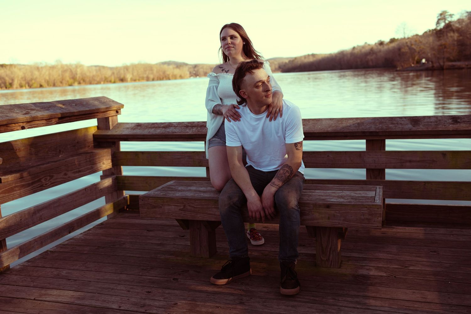 Couple relaxing on a wooden dock by a calm lake, woman standing behind seated man in white shirt.