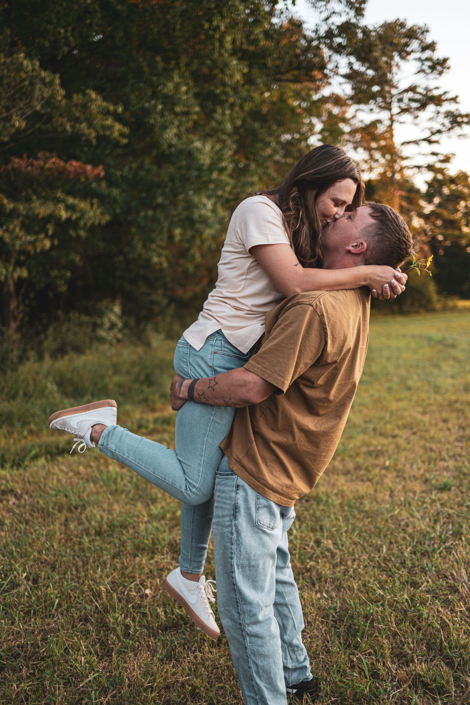 A couple in casual attire embraces playfully in a grassy field during golden hour sunset.