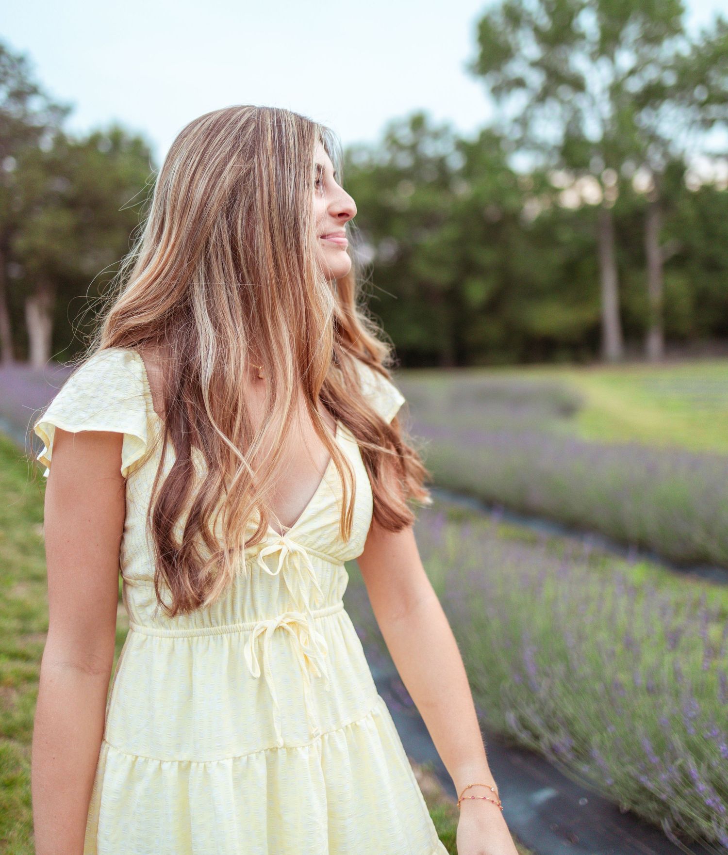 Young woman in yellow dress smiling in lavender field with trees in background.