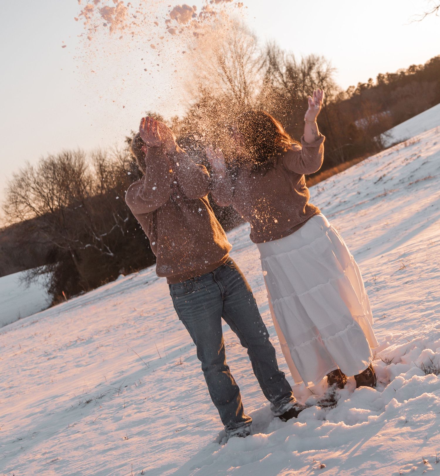 Couple joyfully throwing snow in a snowy winter landscape at golden hour sunset.