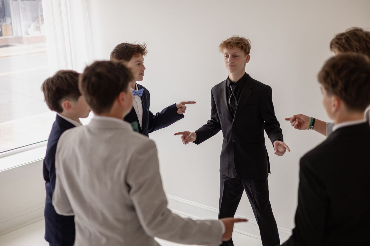 Group of teenage boys in suits pointing fingers at one boy in a dark blazer in a bright white room.