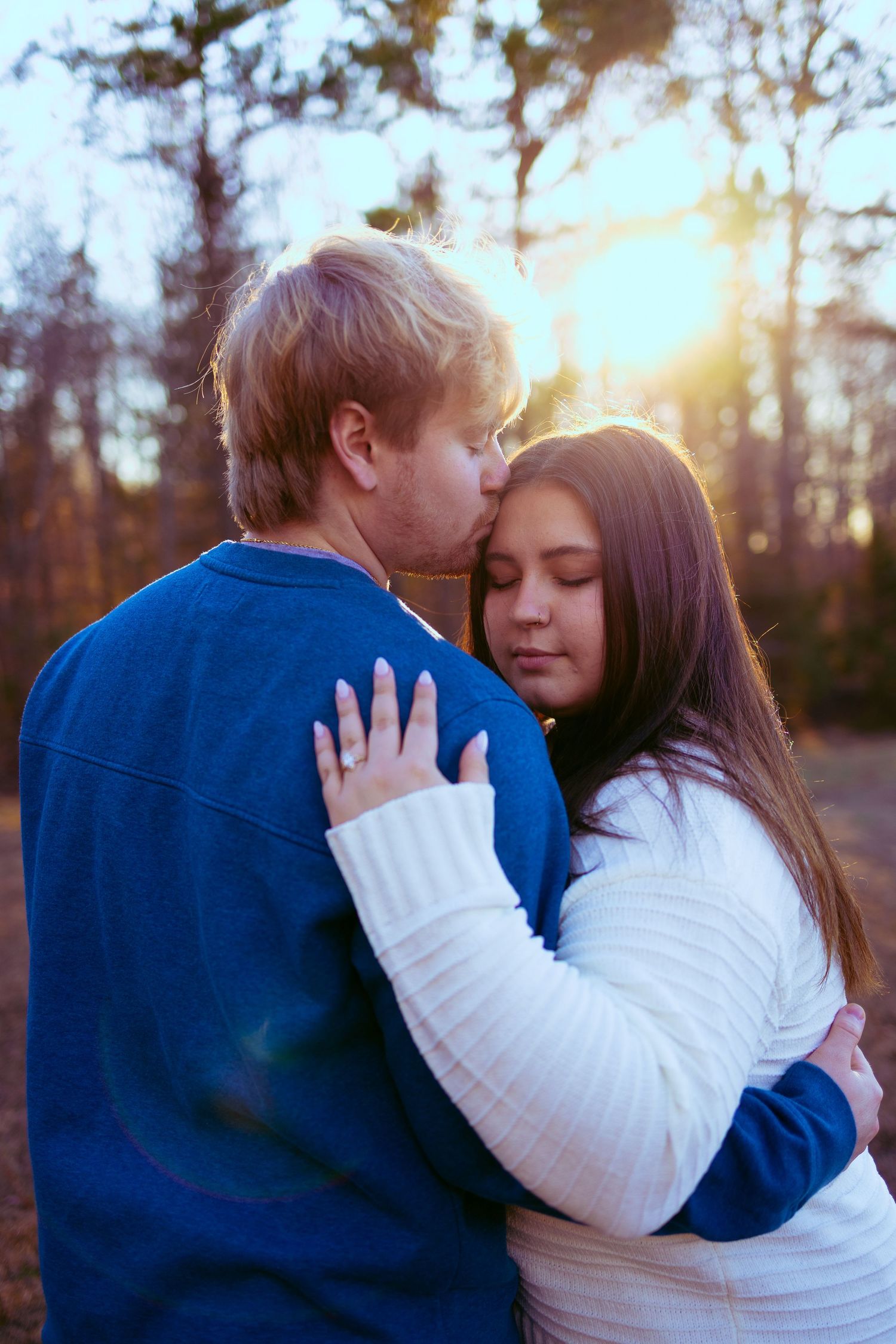 Couple embracing at golden sunset in pine forest, man kissing woman's forehead in romantic outdoor moment.