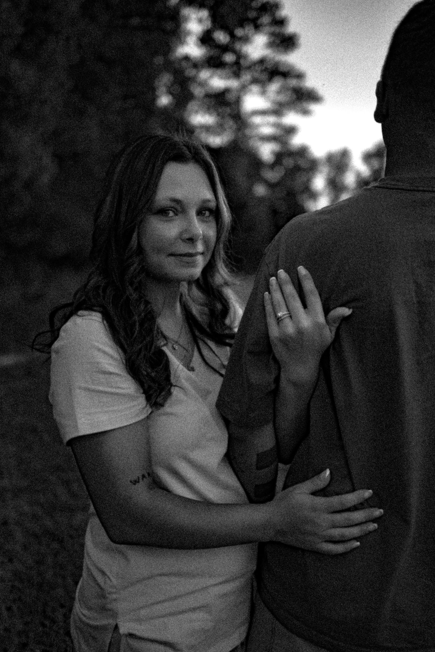 Black and white portrait of a couple embracing outdoors with trees in the background at dusk.