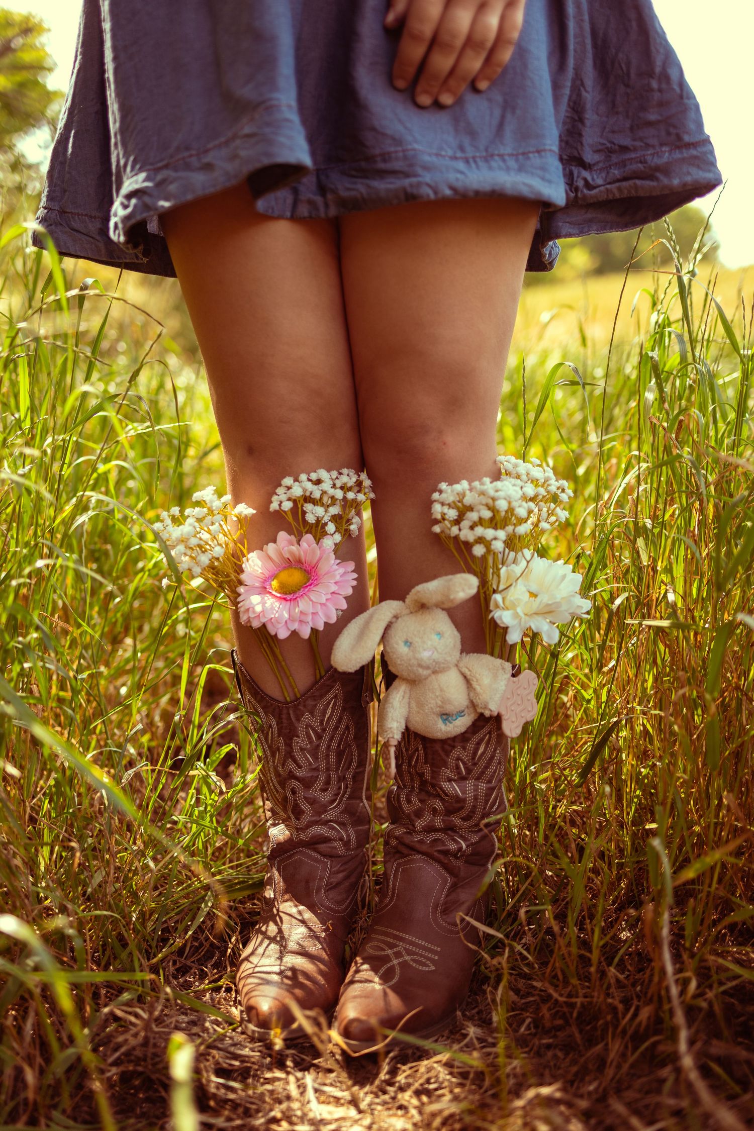 Woman in blue skirt and brown cowboy boots standing in tall grass with colorful flowers tucked into boots.