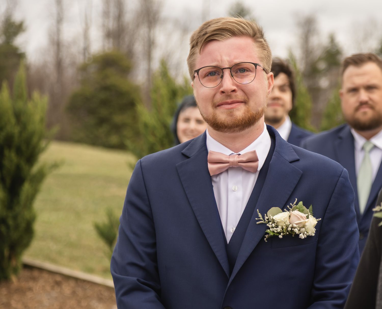 Groomsman in navy suit and pink bow tie poses outdoors with boutonniere.