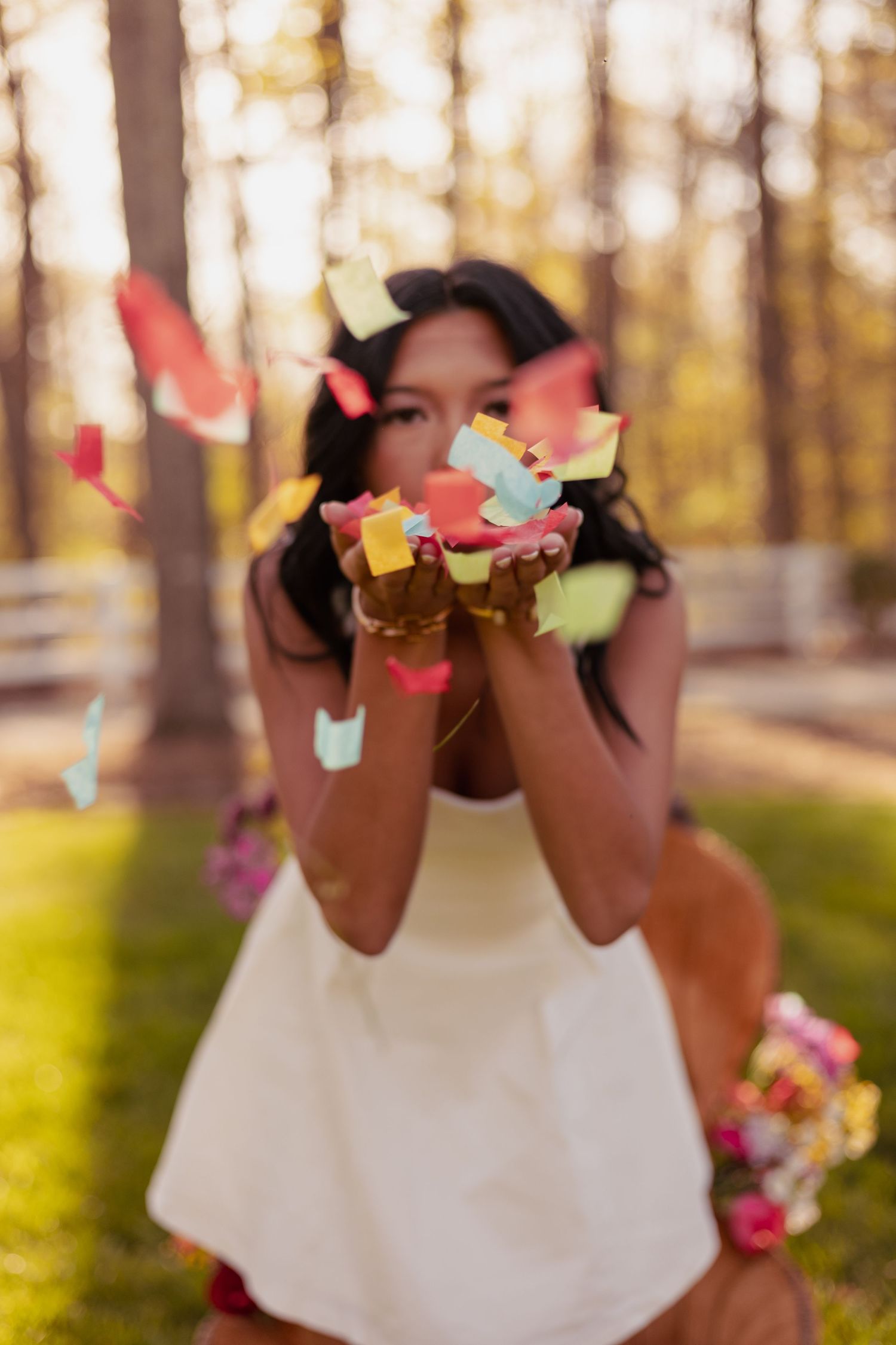 A young woman in a white dress blows colorful confetti from her hands outdoors in a sunlit forest setting.